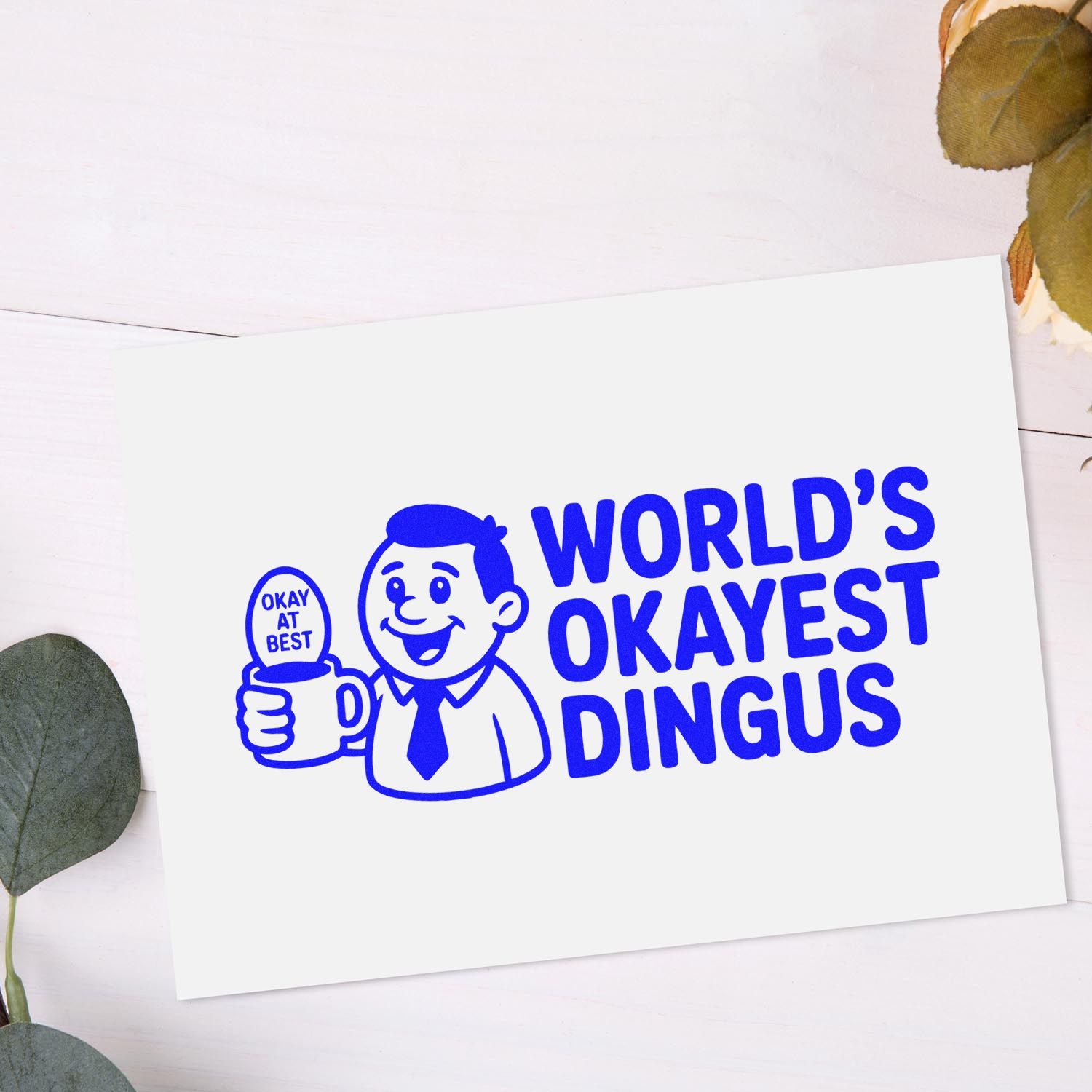 Blue stamp/seal design on white card with smiling man holding an OKAY AT BEST mug and WORLDS OKAYEST DINGUS text.