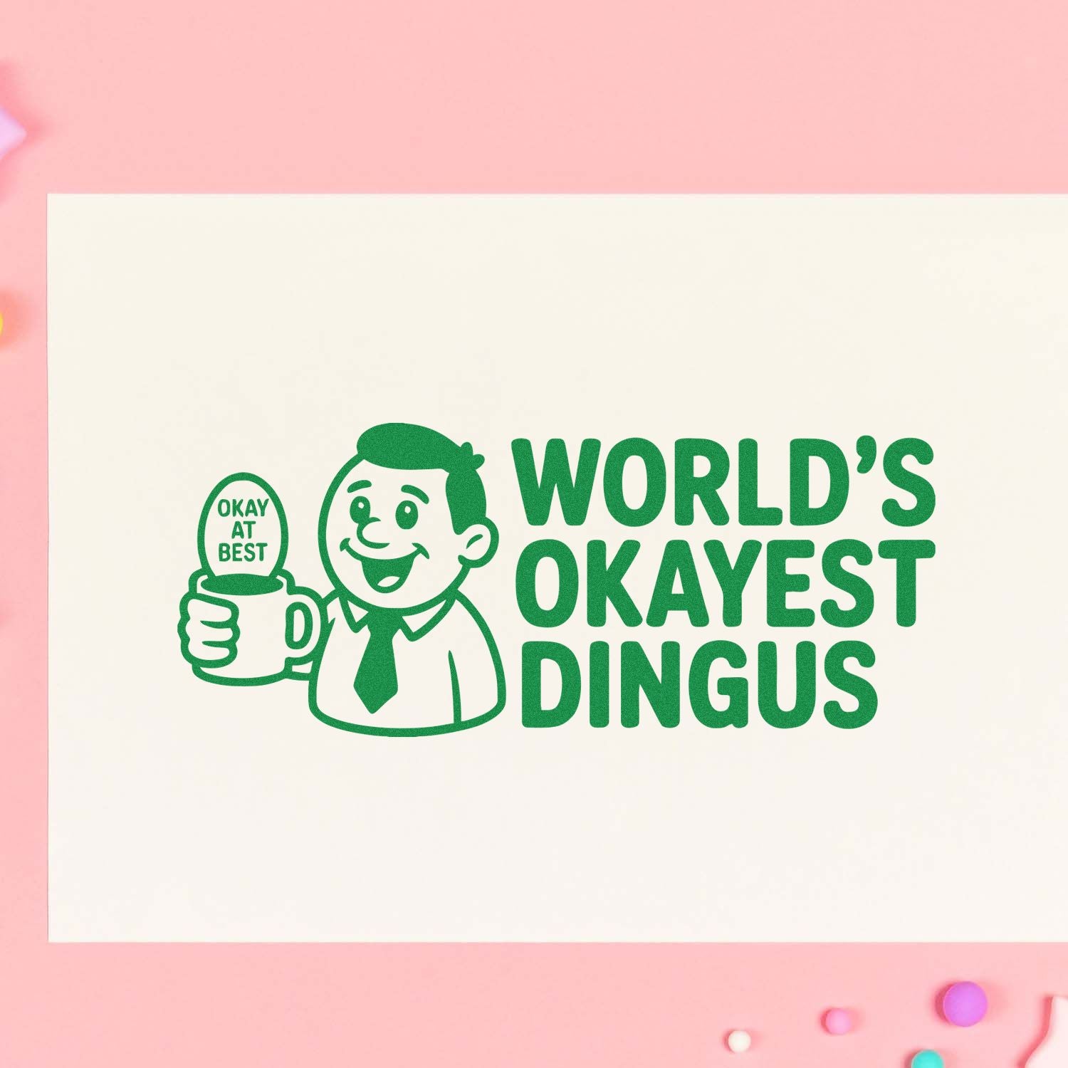 Green stamp design with a smiling man holding an OKAY AT BEST mug and WORLDS OKAYEST DINGUS text on cream paper.