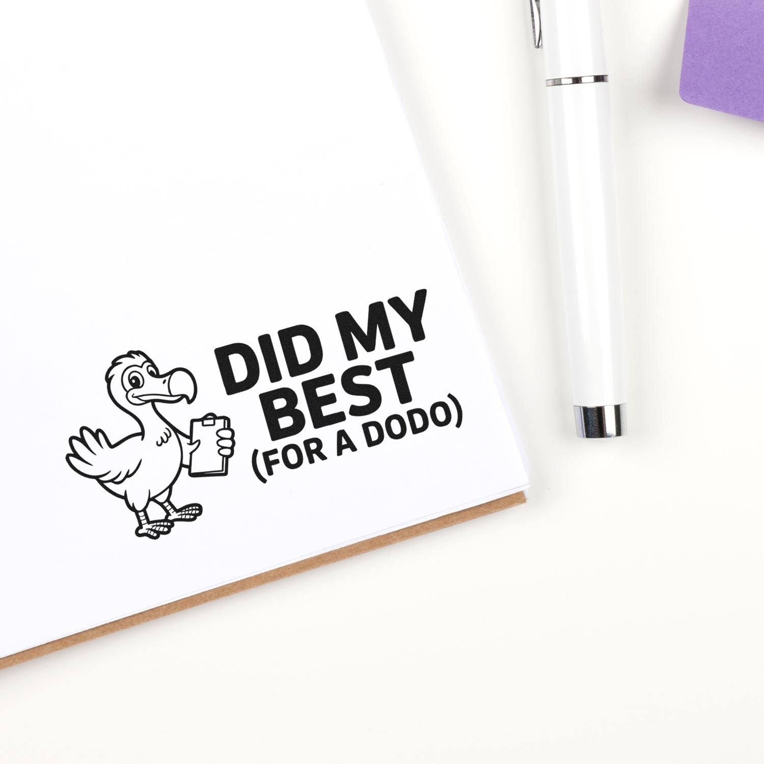 Black custom stamp/seal design with cartoon dodo and DID MY BEST (FOR A DODO) on white paper, beside a white pen and purple note.