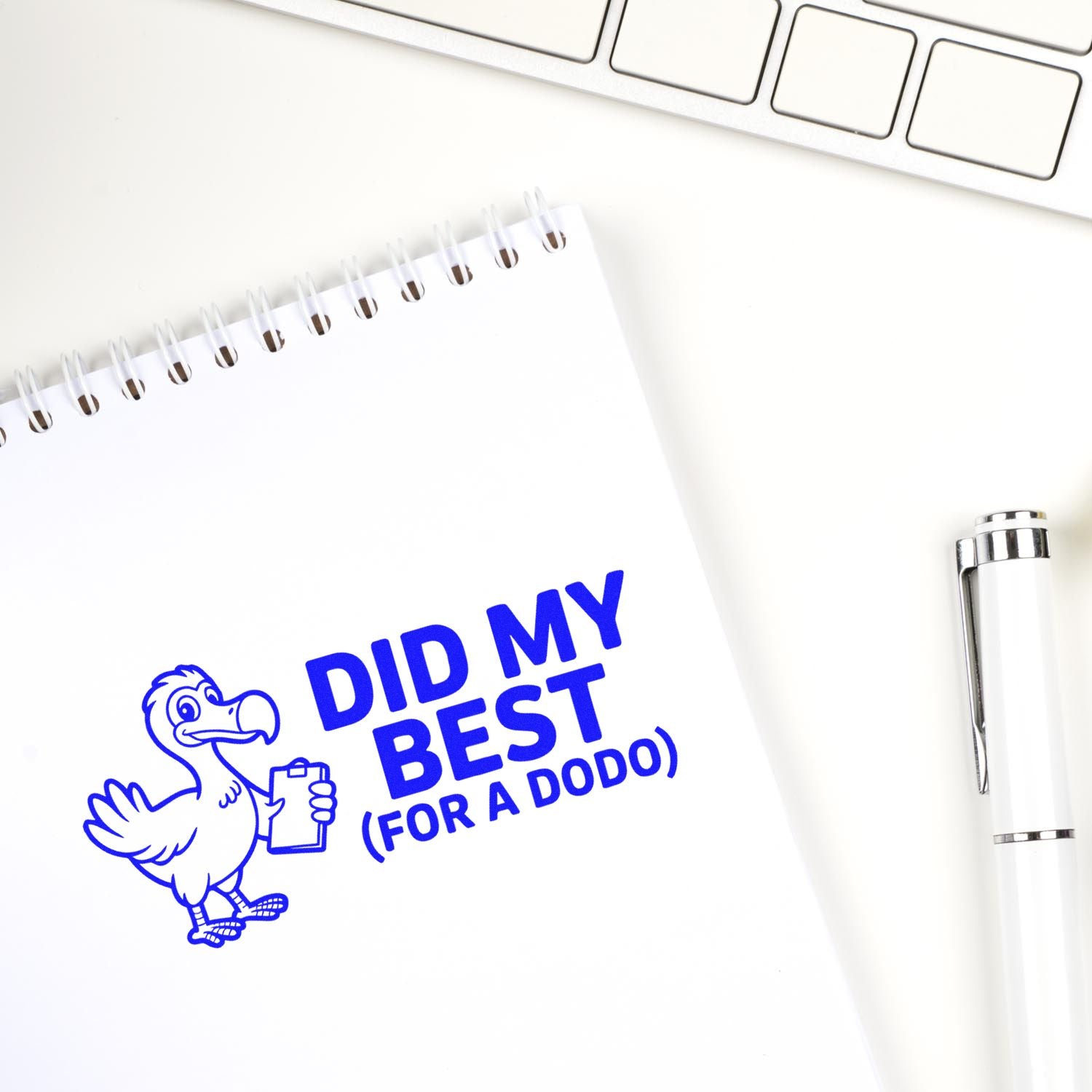 Blue self-inking stamp seal on a white spiral notebook, showing a cartoon bird and Did My Best (For a Dodo) with silver pen.