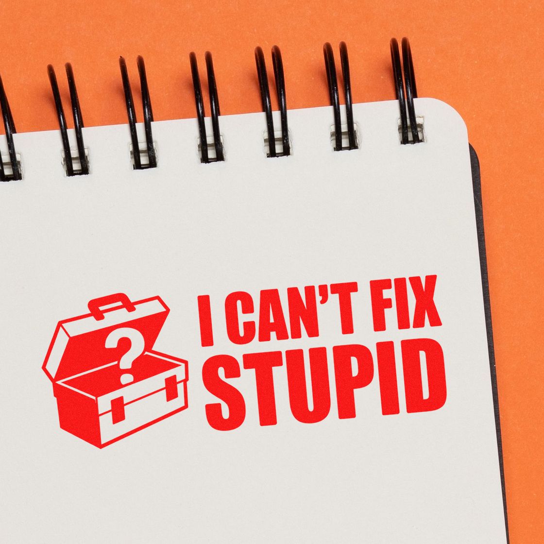 Red stamp-and-seal design reading I CANT FIX STUPID with toolbox icon on a white spiral notebook, orange background.