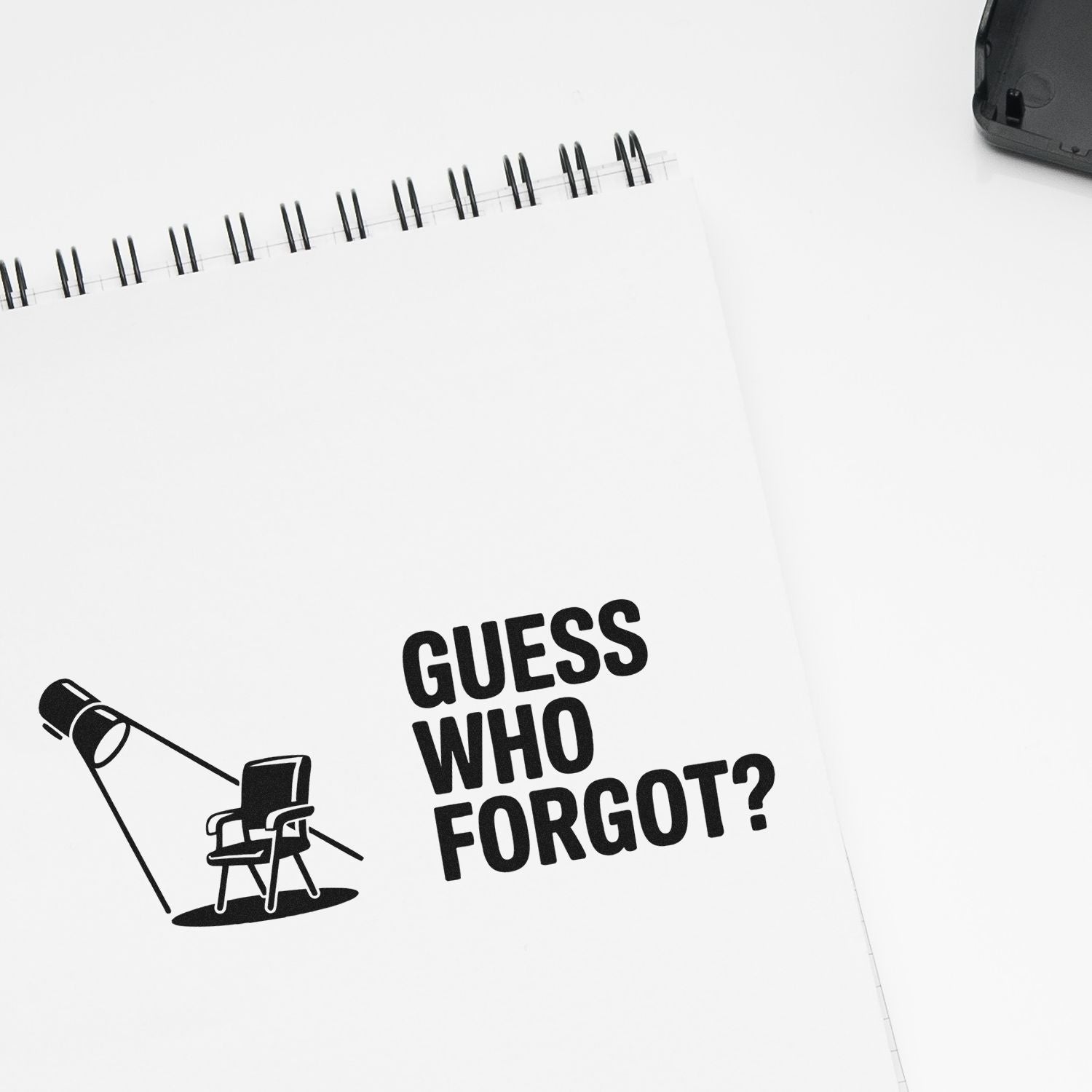 White spiral notepad with black custom stamp and seal design reading Guess Who Forgot? and a desk lamp graphic