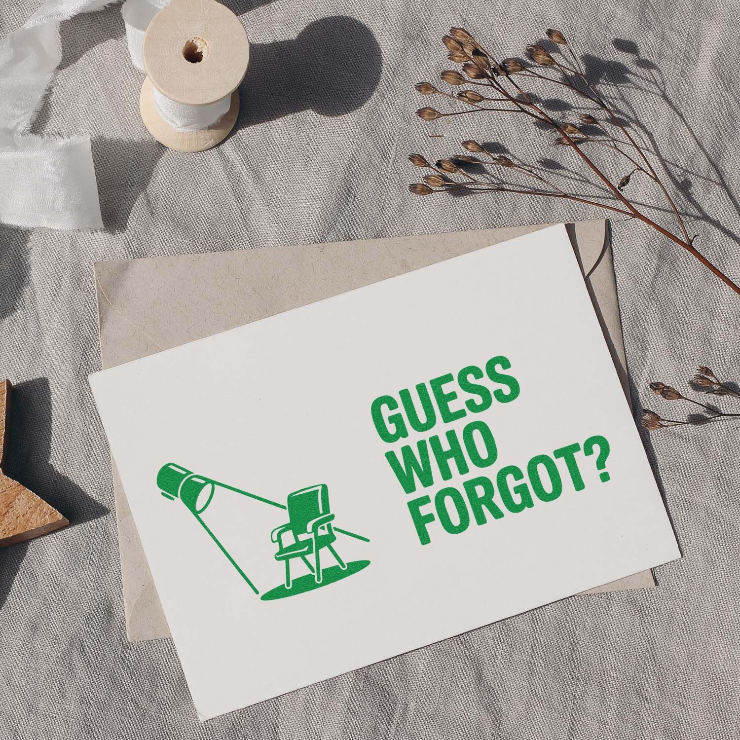 Green stamp and seal card reading GUESS WHO FORGOT? with lamp graphic, on a white card over a beige envelope.