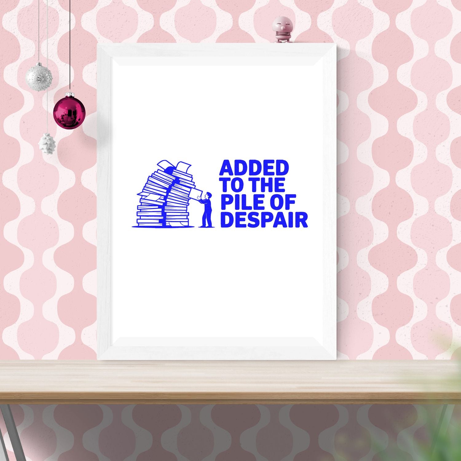 White framed print with blue stamp-style stacked papers and text Added to the Pile of Despair on a pink patterned wall.