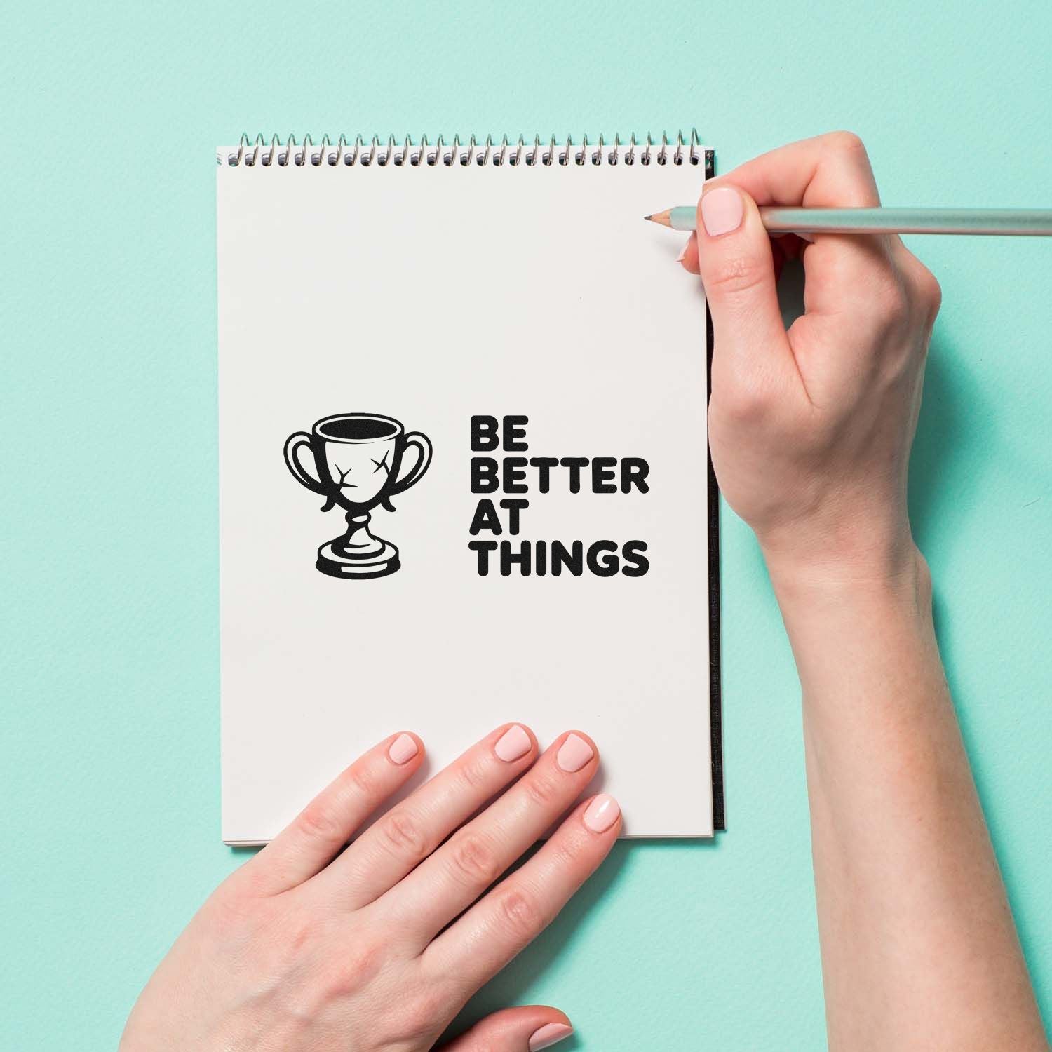 Black trophy stamp and seal reading BE BETTER AT THINGS on white spiral notebook, with hands and pencil on mint background.