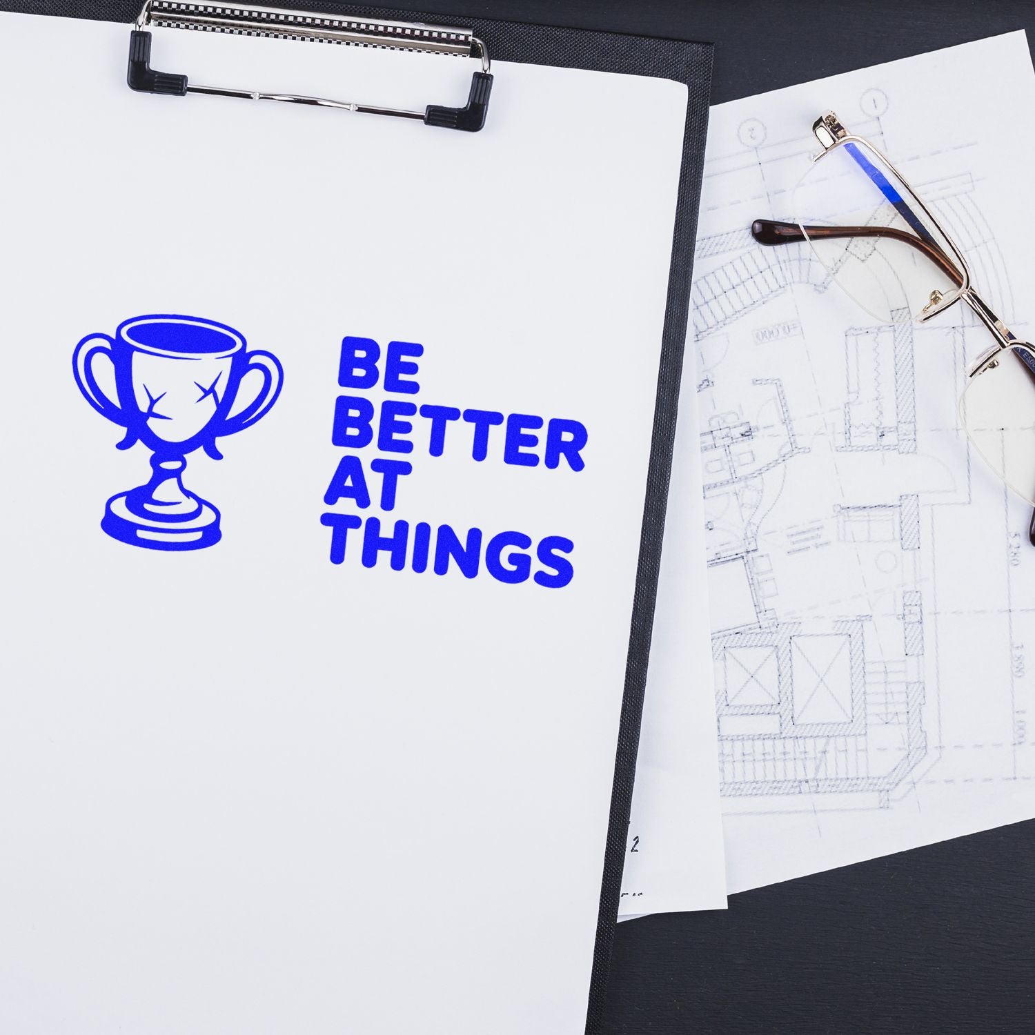 Blue custom stamp and seal trophy reading Be Better at Things on clipboard, beside eyeglasses and architectural plans