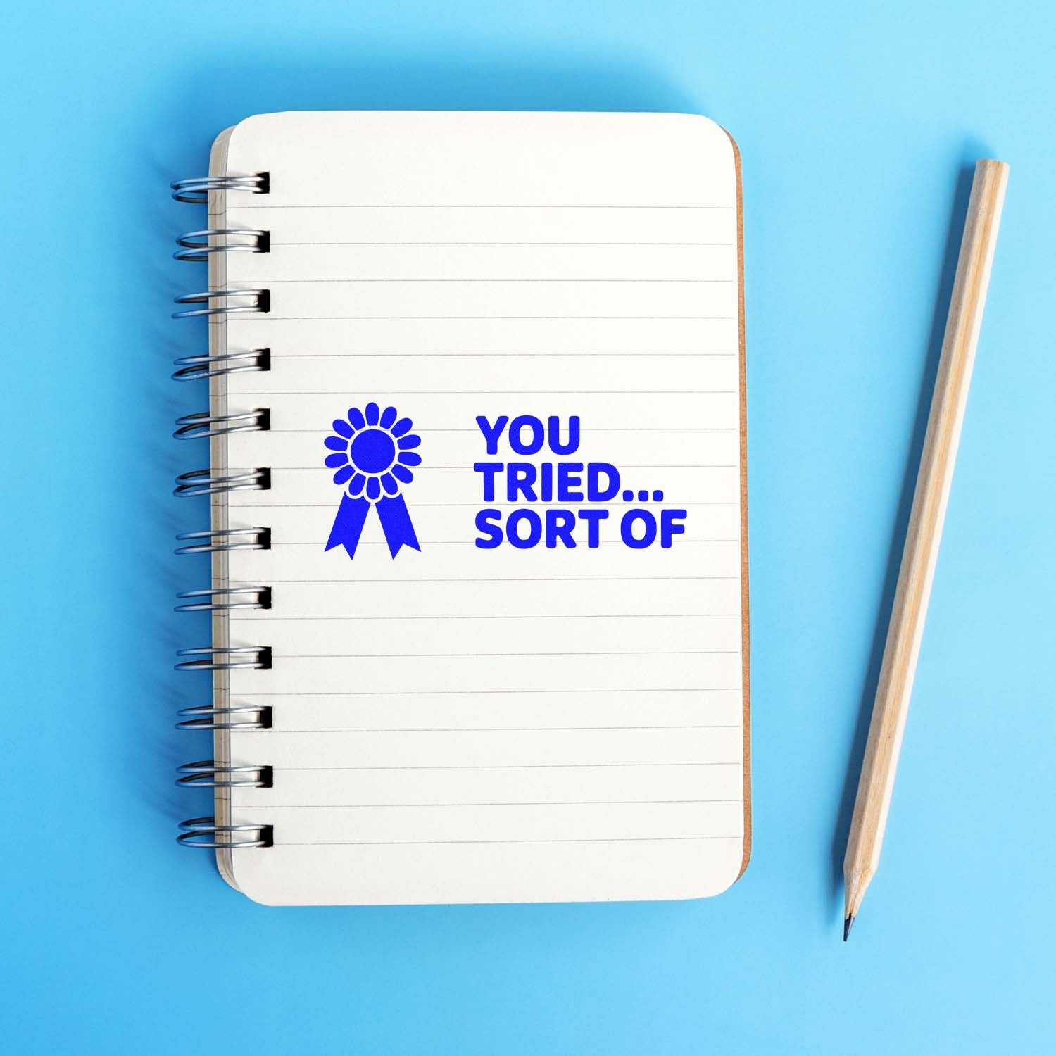 Blue YOU TRIED... SORT OF award seal stamp on a white spiral notebook, with a pencil on a light blue background.