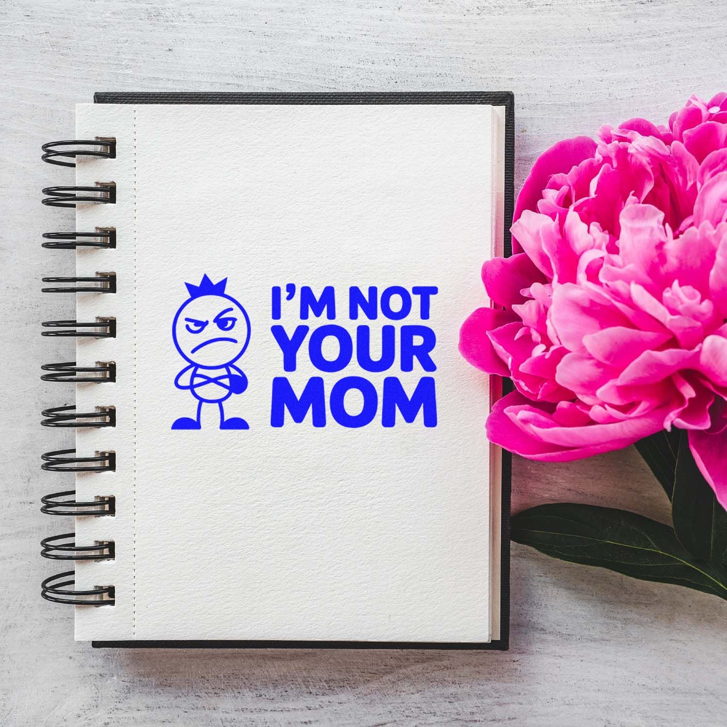 Blue stamp seal reading IM NOT YOUR MOM on a white spiral notebook beside bright pink peonies.