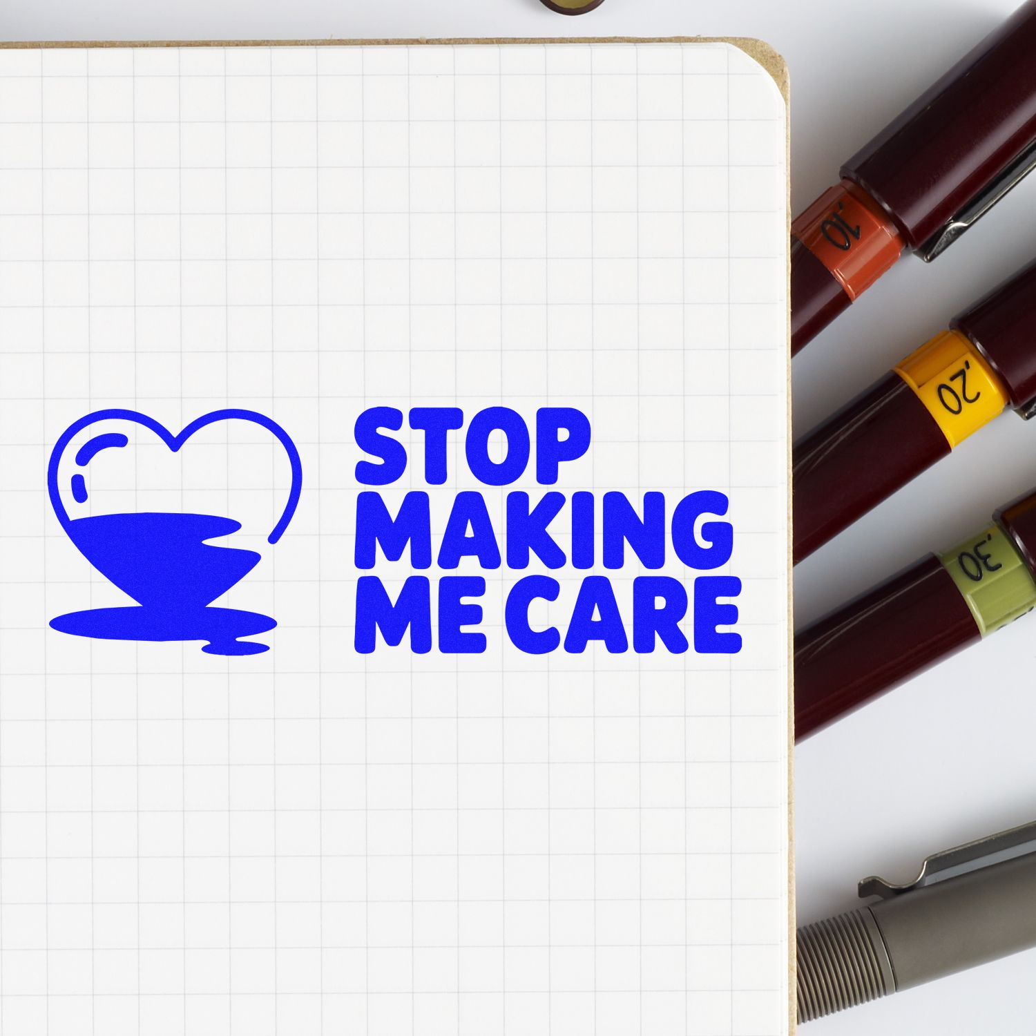 Blue STOP MAKING ME CARE stamp with heart icon on grid notebook, beside maroon seal markers with orange, yellow, and green caps.