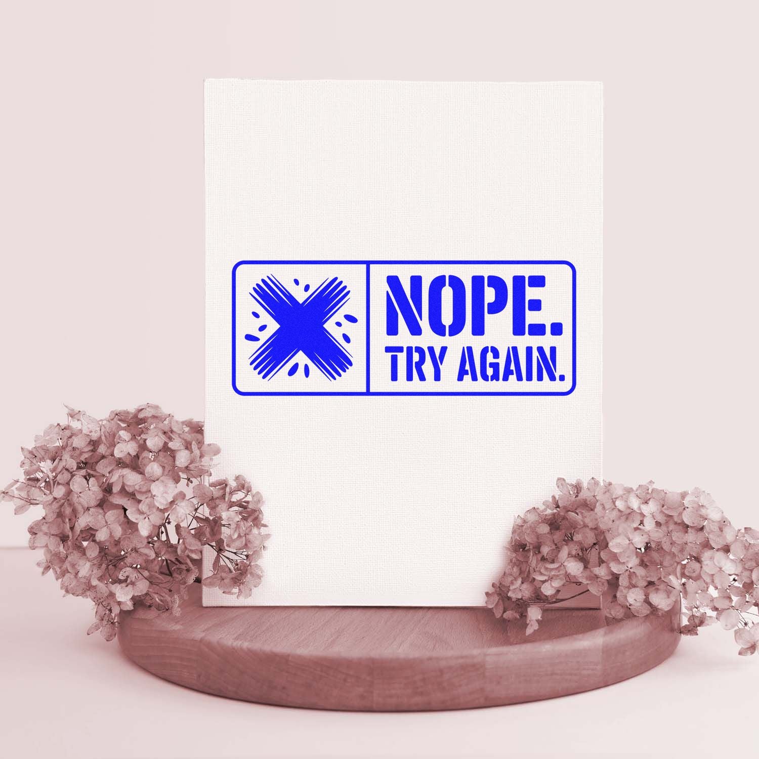 Blue stamp-style seal reading NOPE. TRY AGAIN. on a white card, framed by pink flowers on a wooden base.