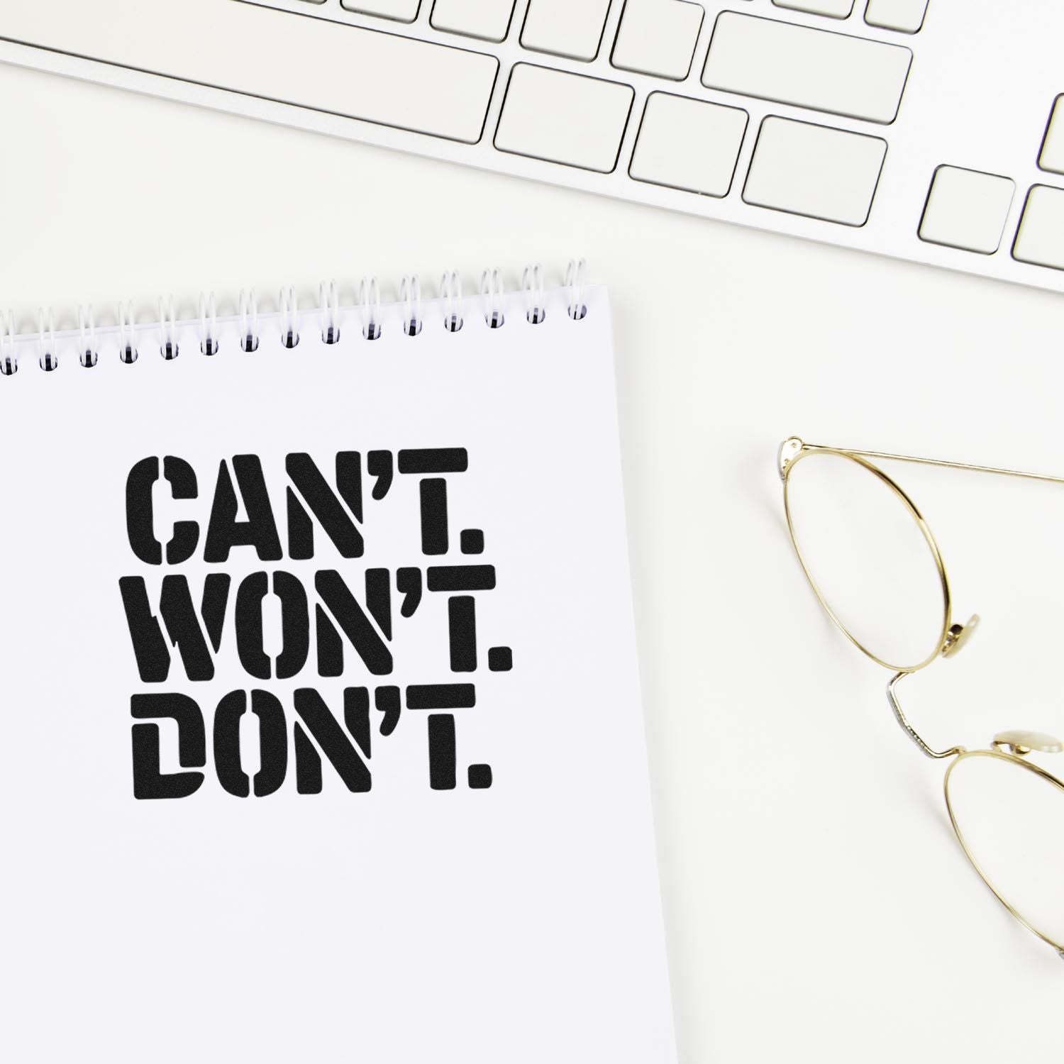 Bold black CANT WONT DONT custom stamp on white spiral notebook, with gold glasses and keyboard on a white desk.