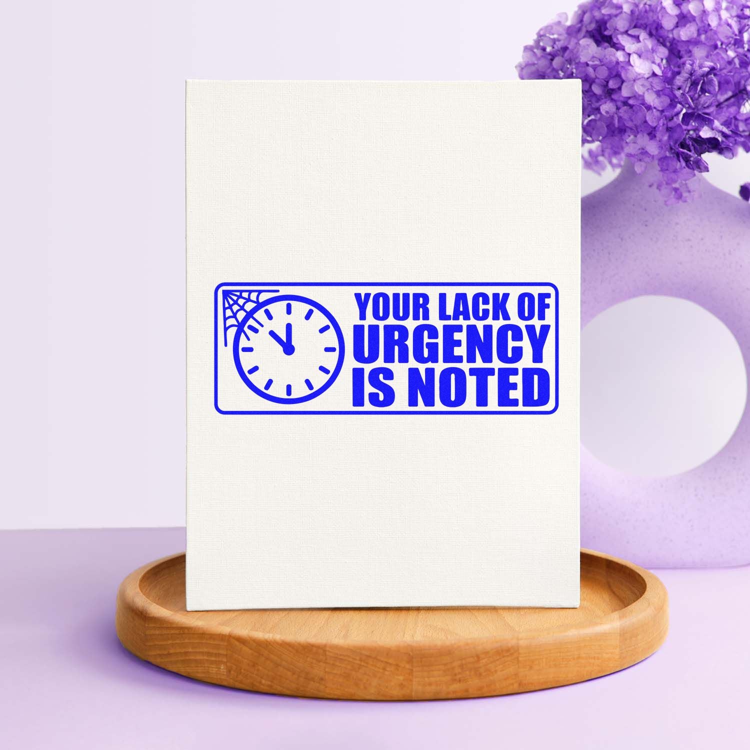Blue rubber stamp on white card reading Your Lack of Urgency Is Noted, with clock and spiderweb, on a wooden display base.