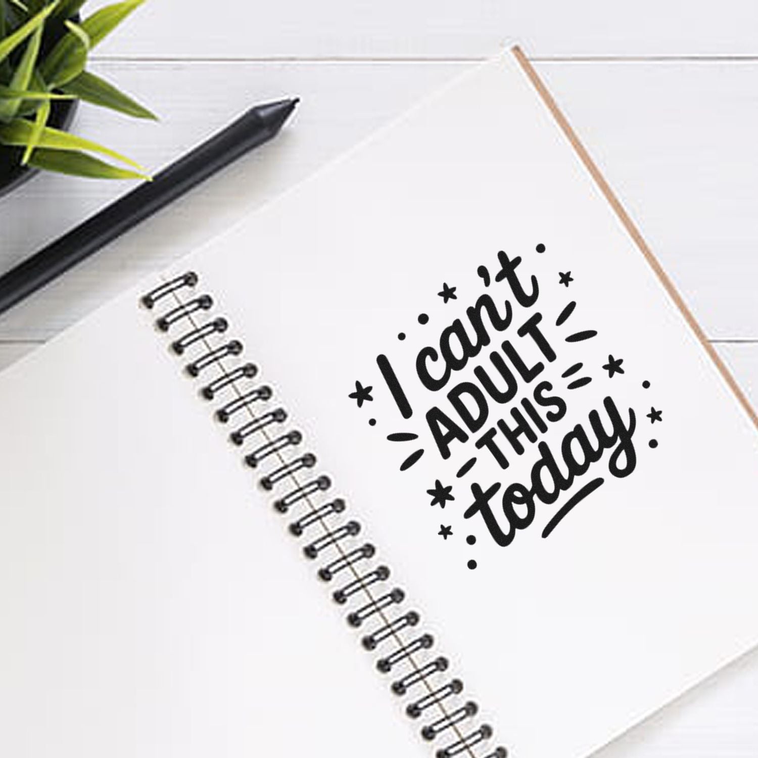 Black stamp seal reading I cant adult today on a white spiral notebook, with a black pen and green plant nearby.