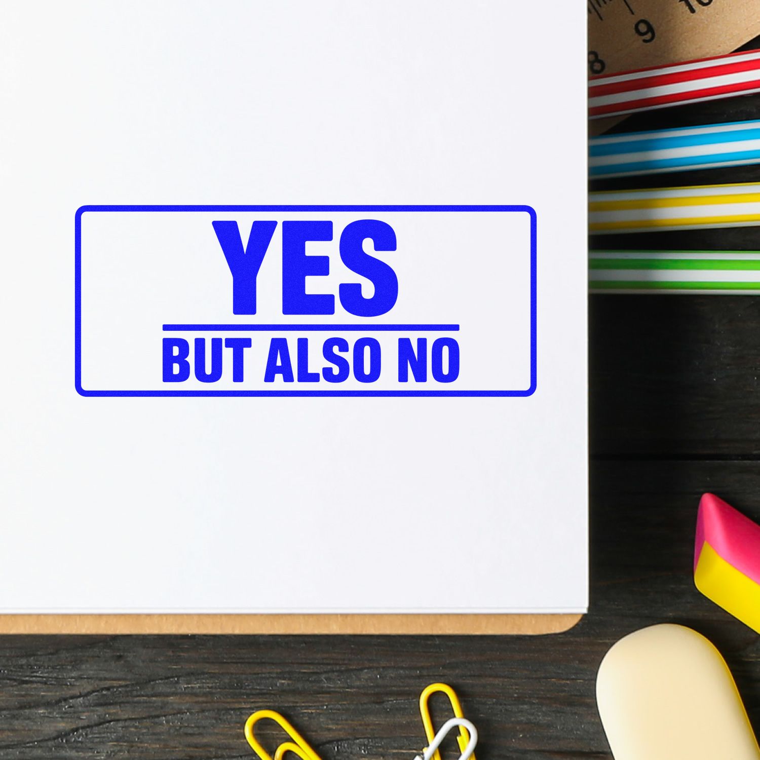 Blue rectangular rubber stamp reading YES BUT ALSO NO on white paper, with colorful stationery tools nearby.