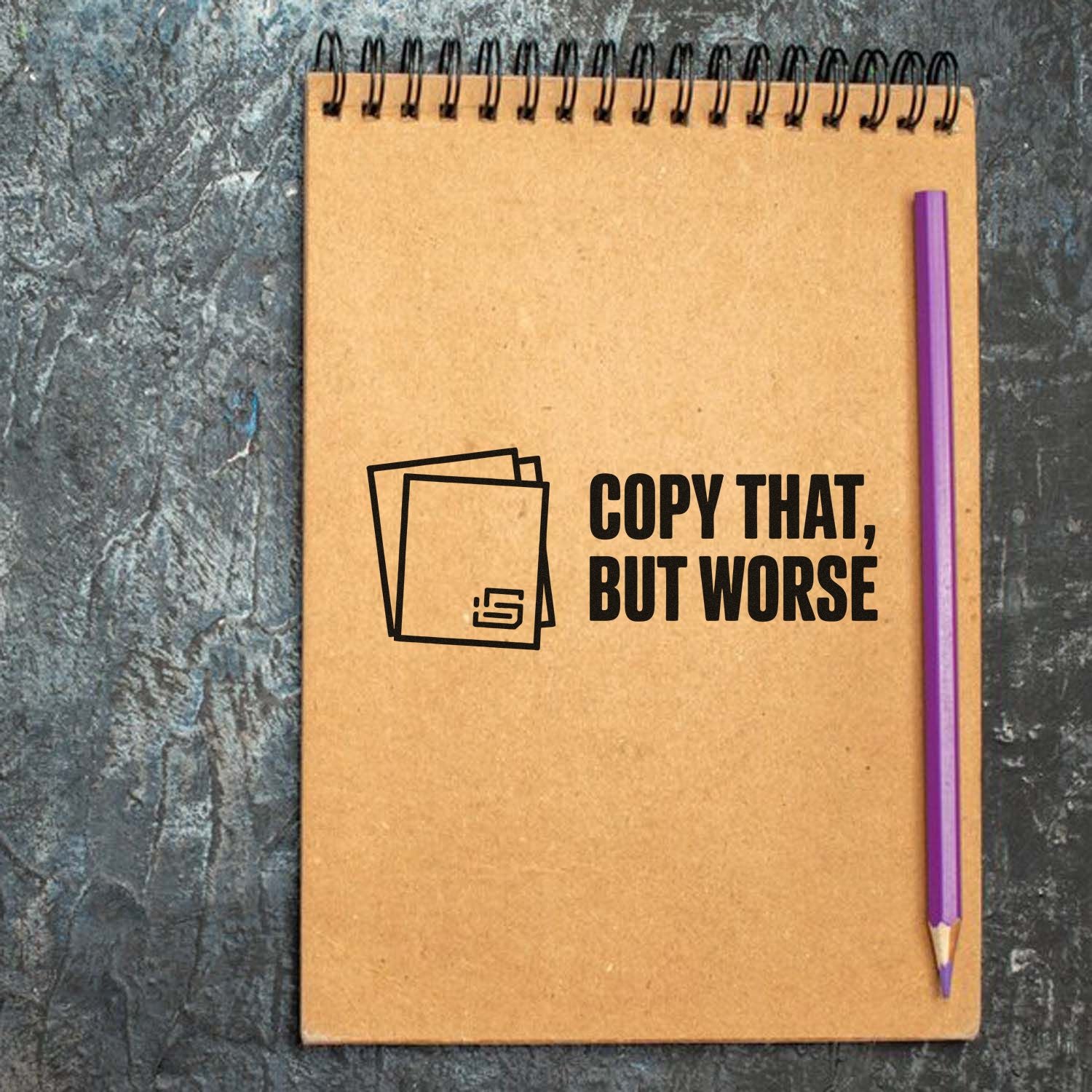 Kraft spiral notebook with black stamp-and-seal logo and Copy That, But Worse text, plus a purple pencil.