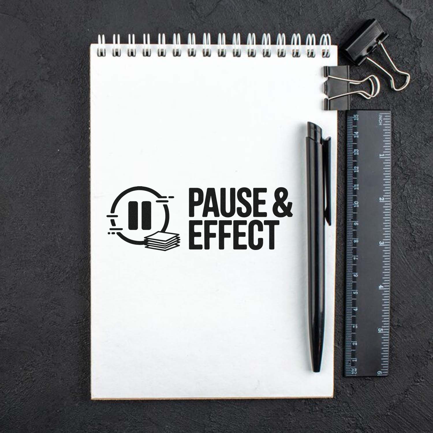 Black stamp seal reading Pause & Effect on a white spiral notebook, with pen, ruler, and binder clips on a dark background.