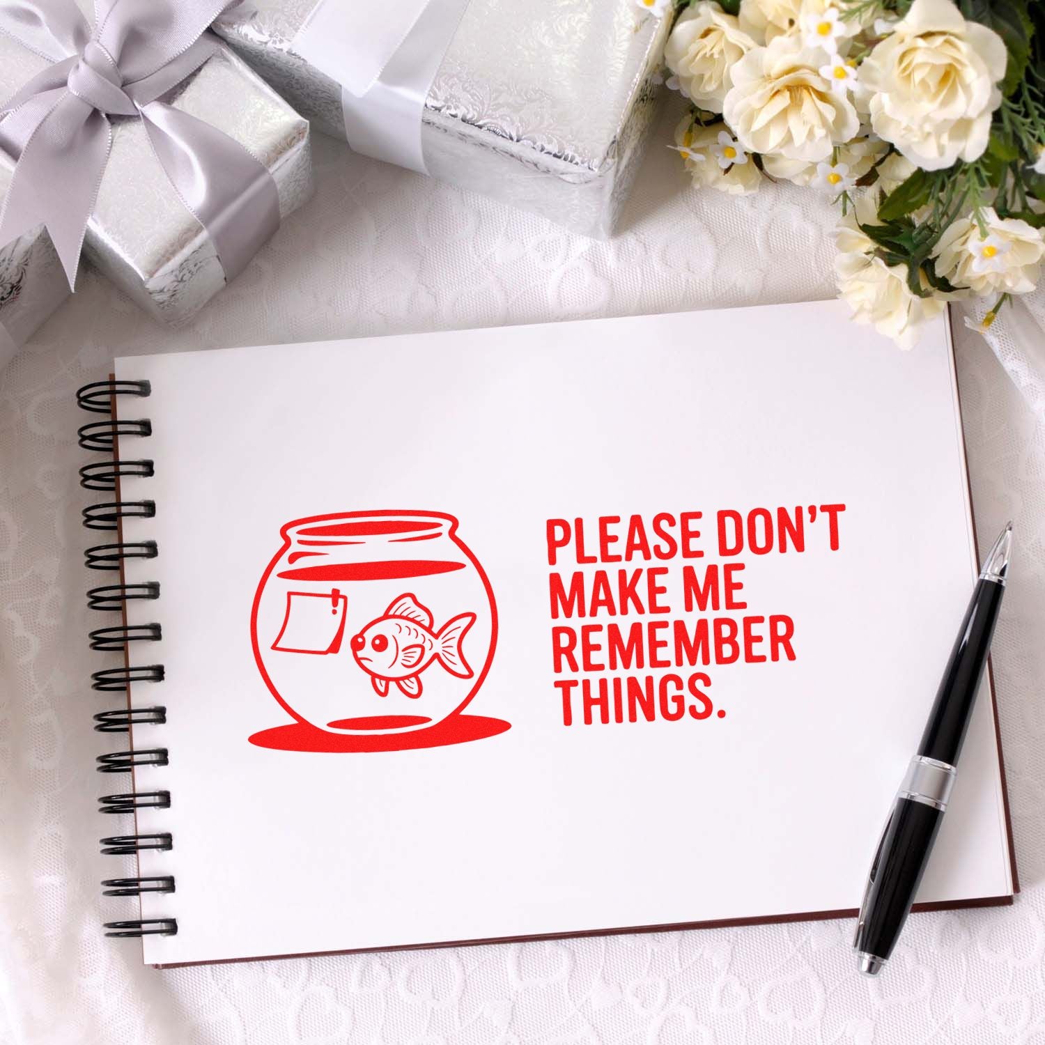 Red stamp and seal design on white spiral notebook reads Please dont make me remember things, with goldfish bowl, gifts, flowers, and pen.