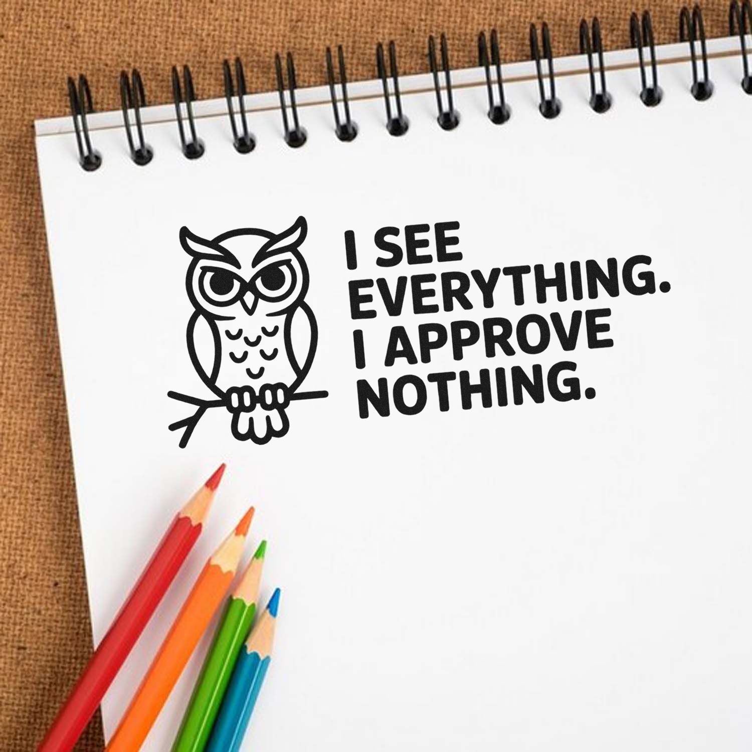 Black owl stamp and seal with bold text I SEE EVERYTHING. I APPROVE NOTHING. on white spiral notebook, colored pencils.
