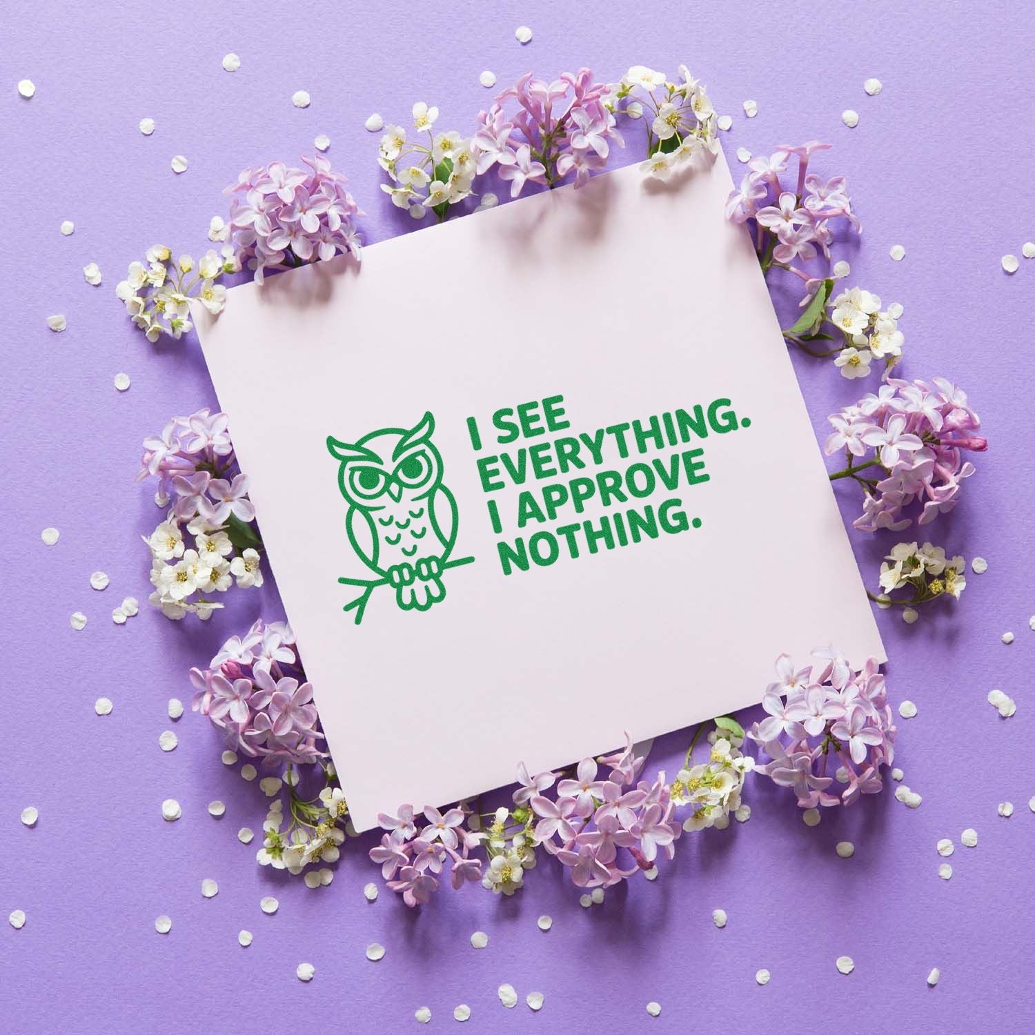 Green owl rubber stamp and seal with I see everything. I approve nothing. on white card, surrounded by lilac flowers on purple background.