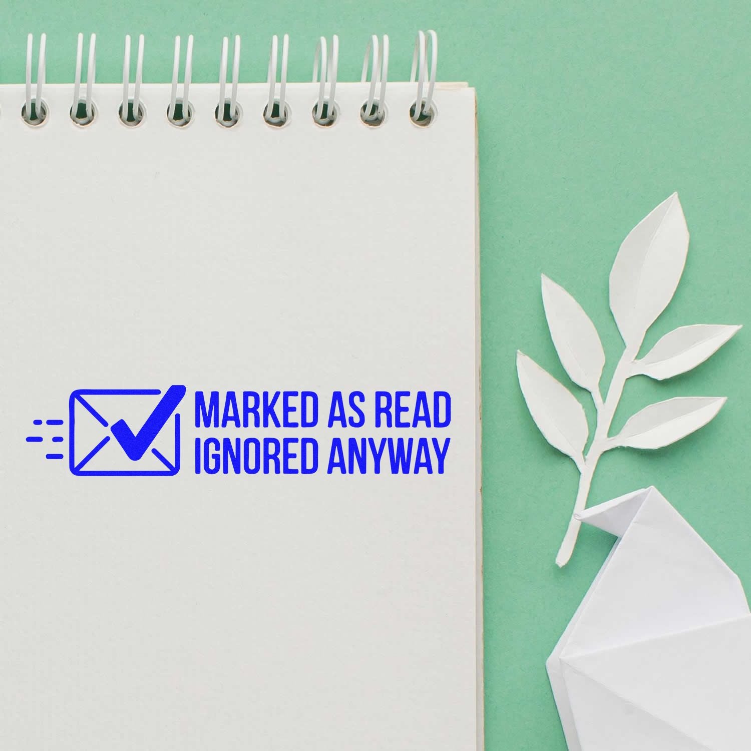 Blue self-inking stamp/seal reading Marked as Read Ignored Anyway on white notebook, with mint green background and white paper leaves.