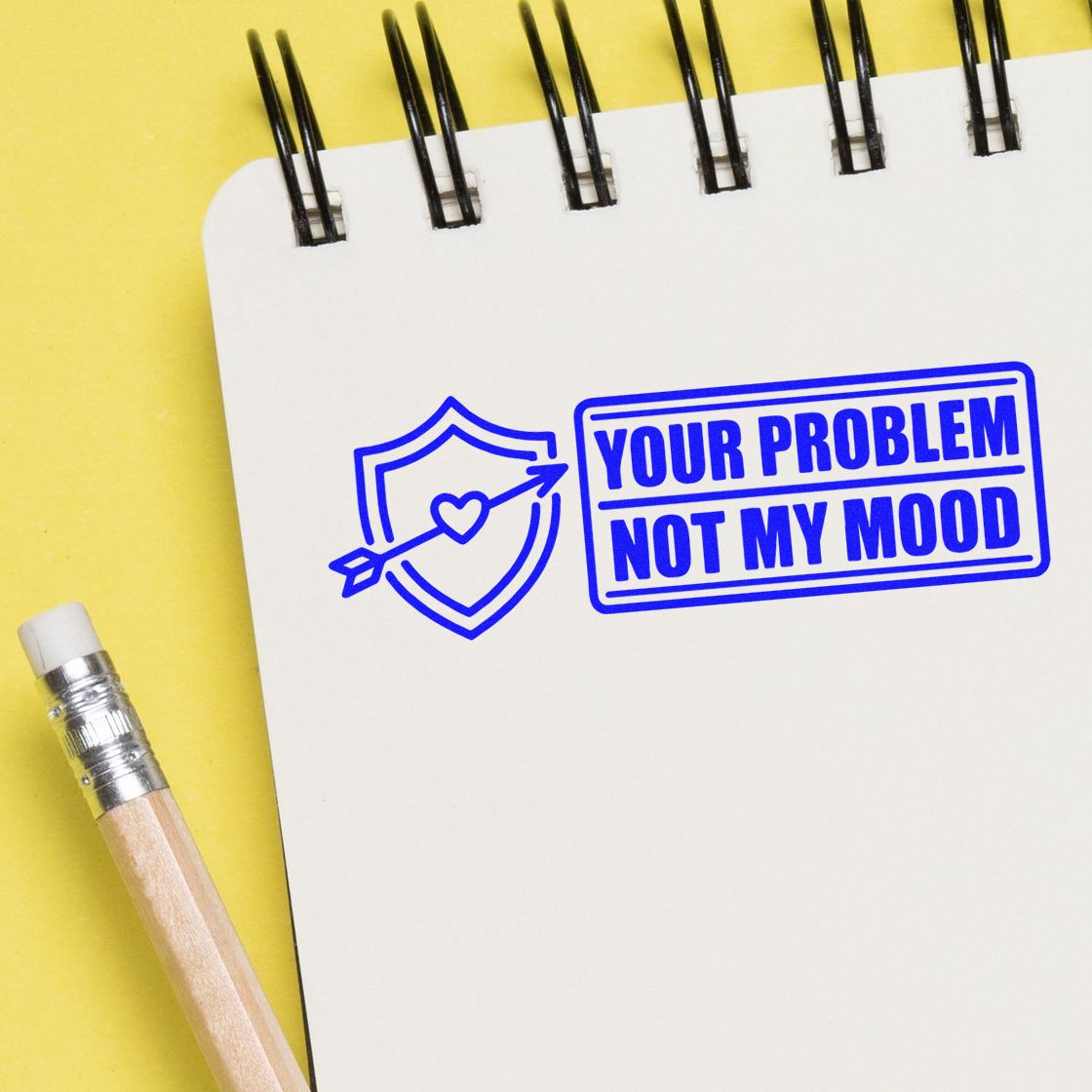 Blue rubber stamp seal on white spiral notepad, saying Your problem not my mood, with shield and heart arrow on yellow background.