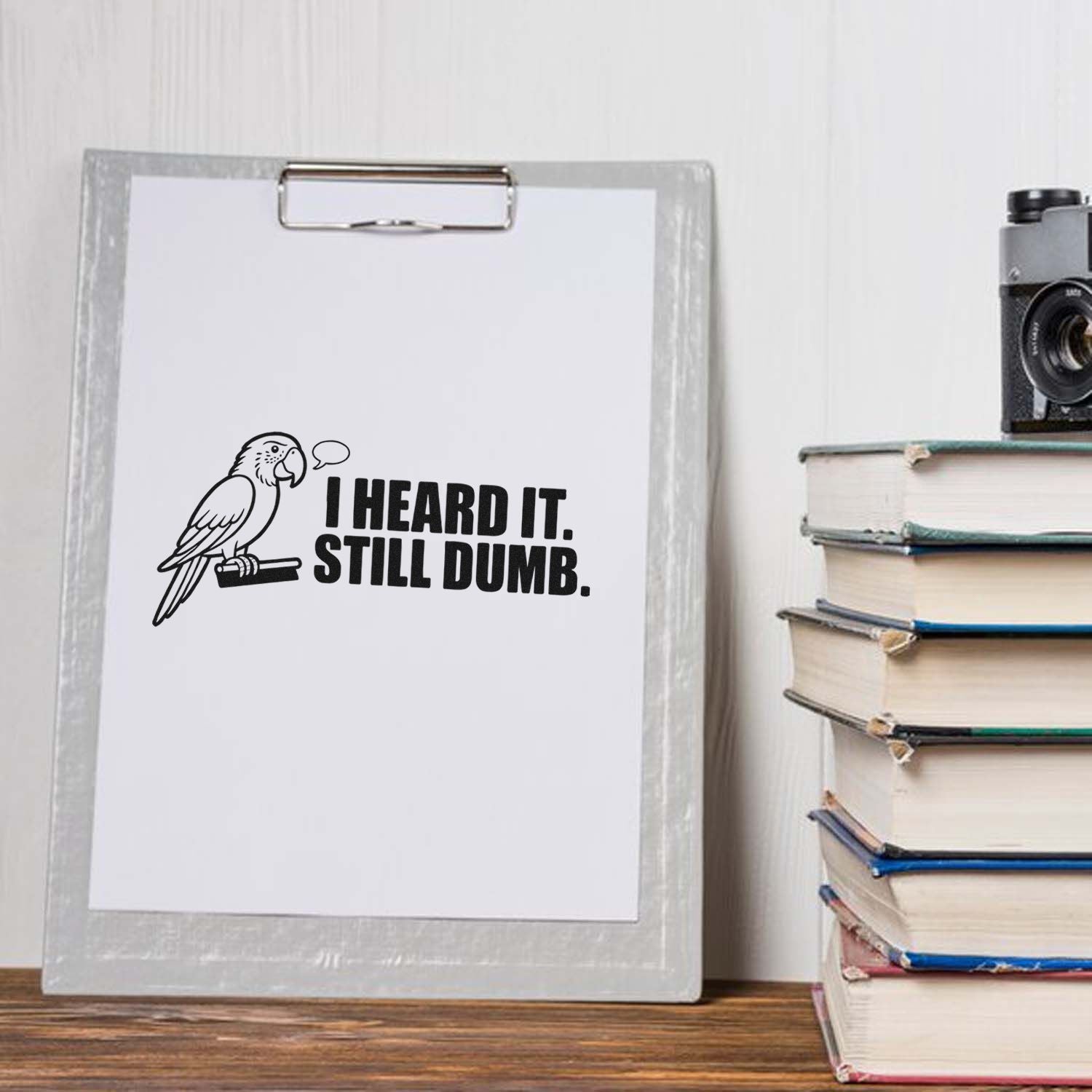 Black parrot rubber stamp design with bold text I HEARD IT. STILL DUMB. on white clipboard beside stacked books.