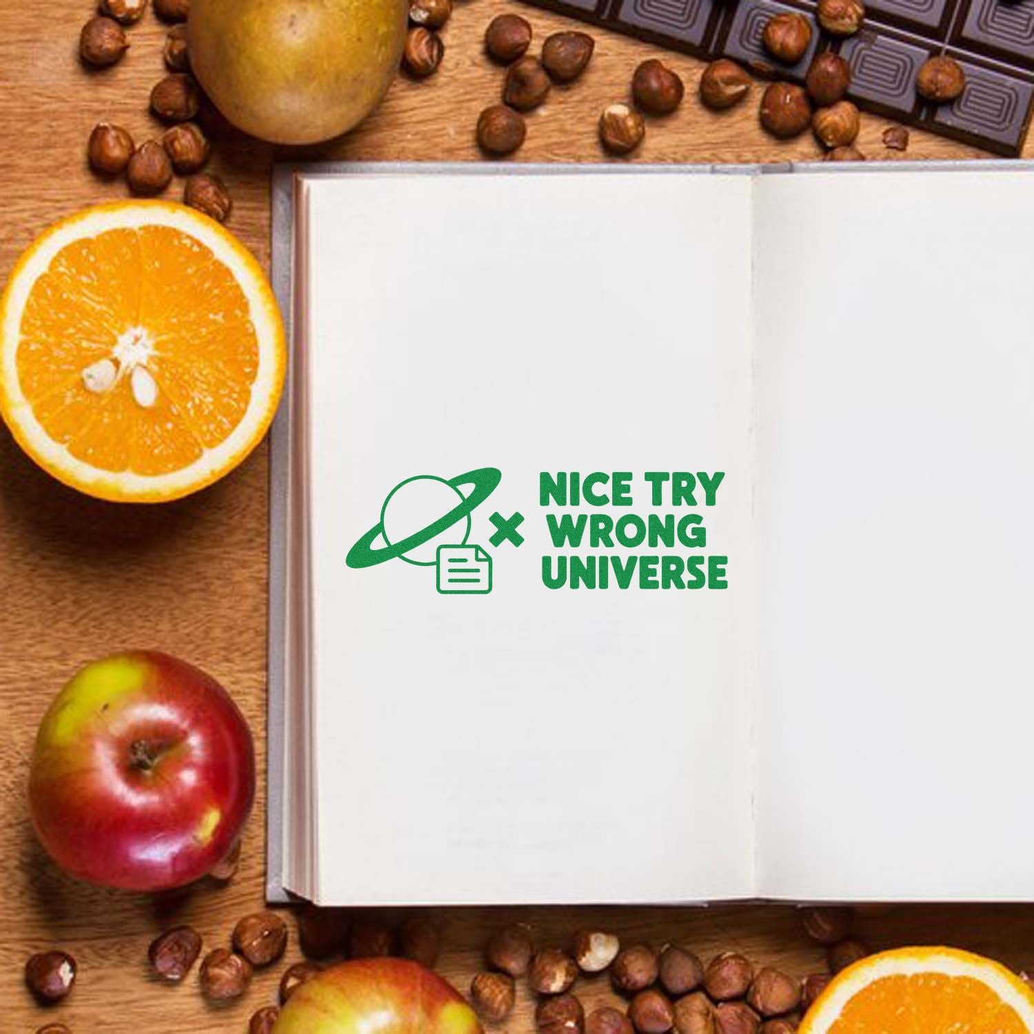 Green rubber stamp seal reading Nice Try Wrong Universe on an open notebook, with oranges, apples, and coffee beans.