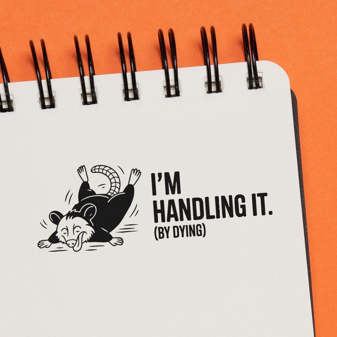 White spiral notebook with a black opossum stamp design and bold Im handling it (by dying) text on an orange background.