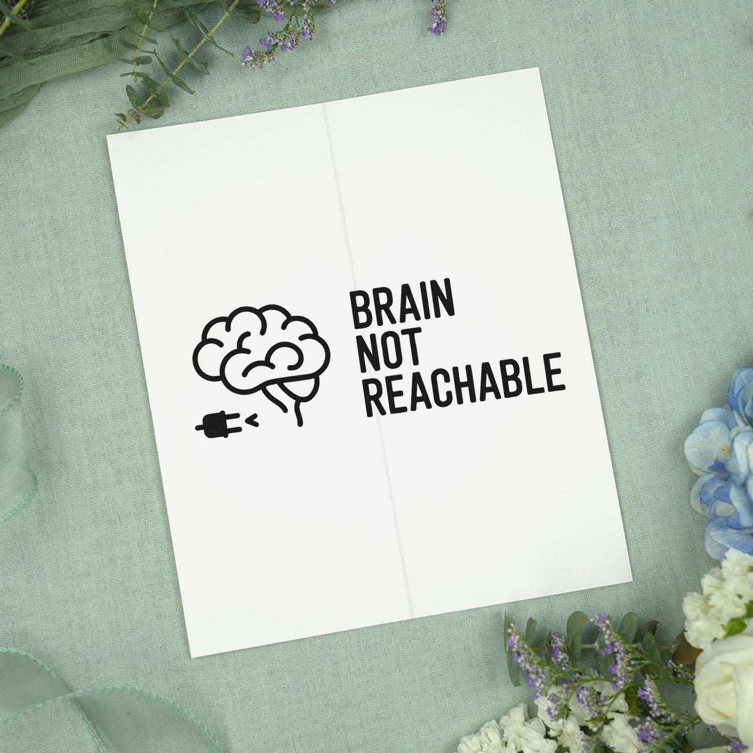 Black brain stamp seal with unplugged icon and BRAIN NOT REACHABLE on white card, styled on mint fabric with flowers.