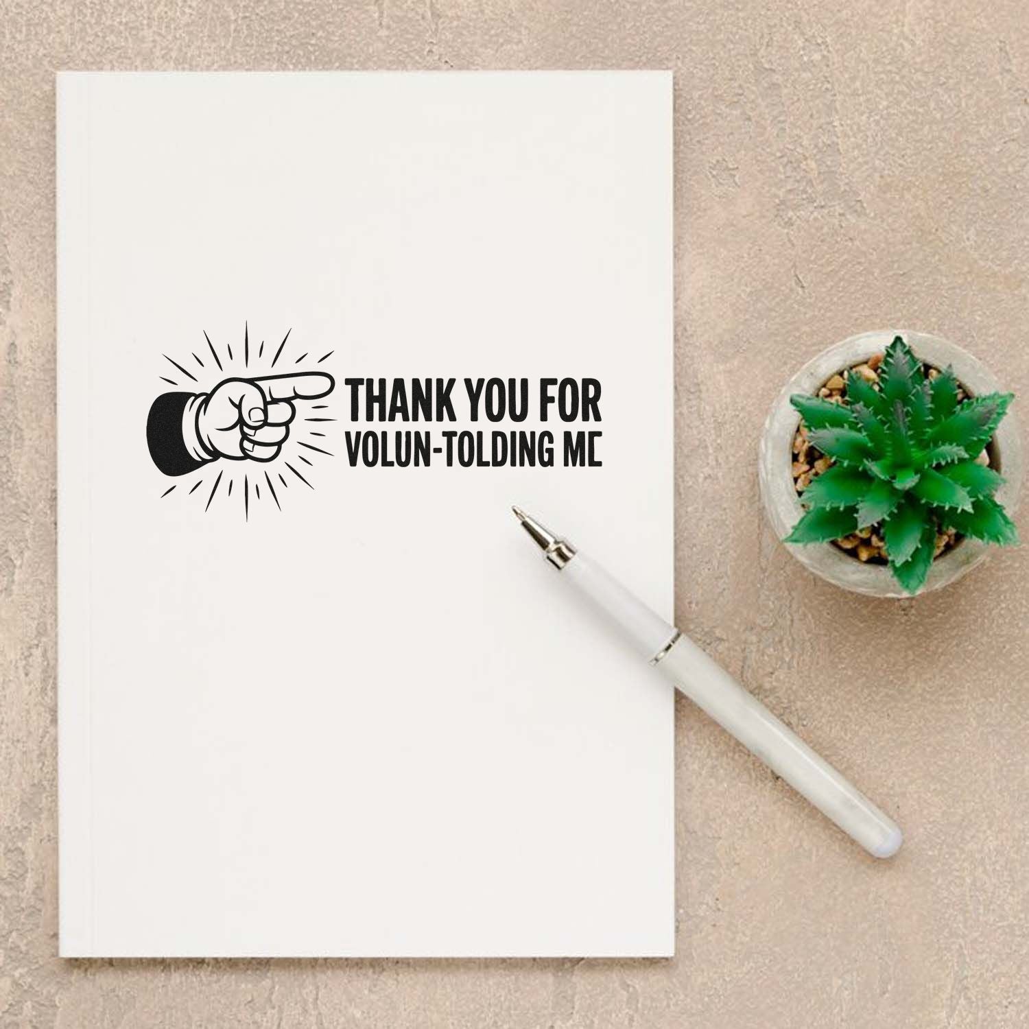 White notebook with black custom thank-you stamp, pointing hand graphic, white pen, and green succulent on beige background.