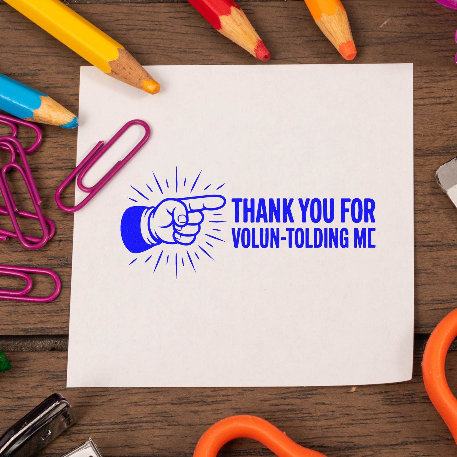 Blue rubber stamp seal with pointing hand, reading "Thank You for Volun-Tolding Me," on white paper with colorful pencils.