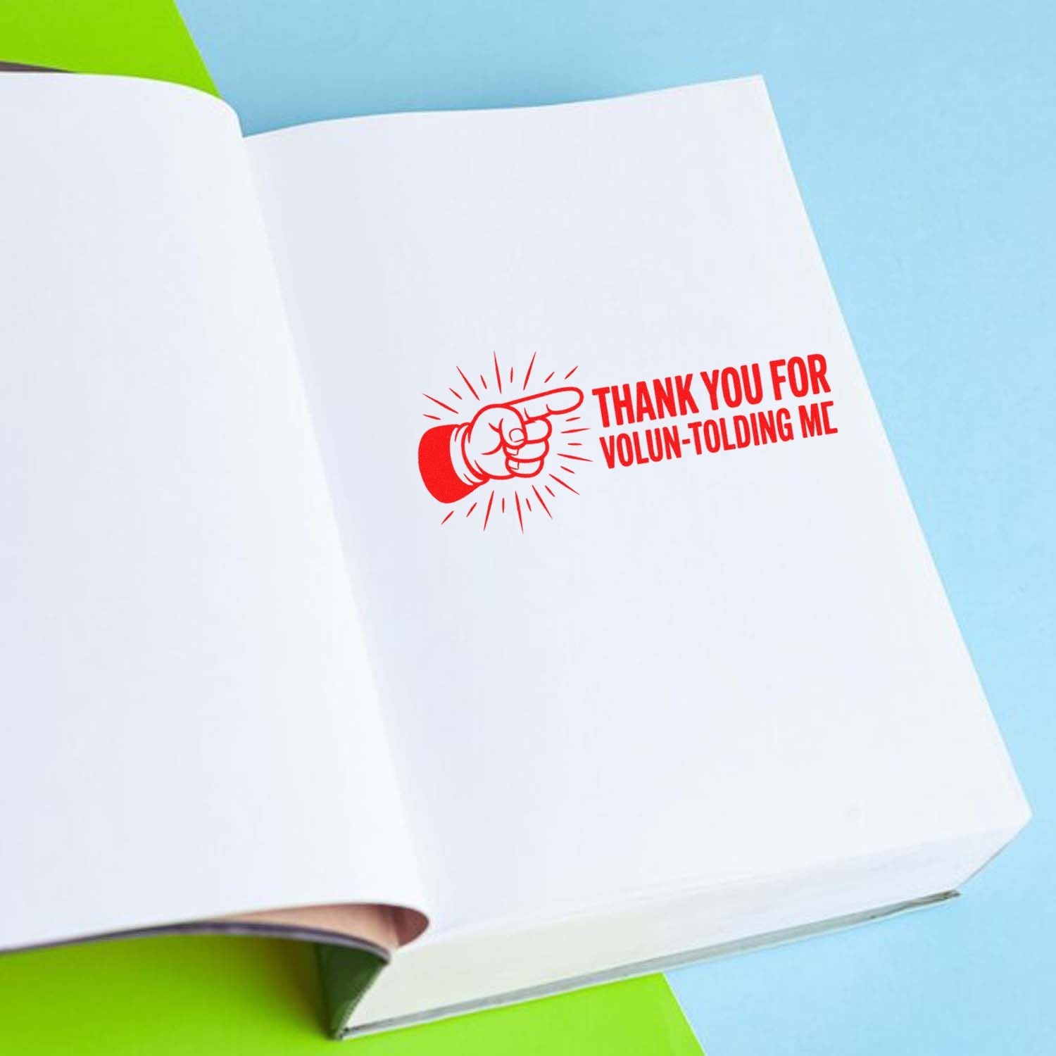 Open white notebook with red pointing-hand rubber stamp/seal reading Thank you for volun-tolding me on blue and green background.