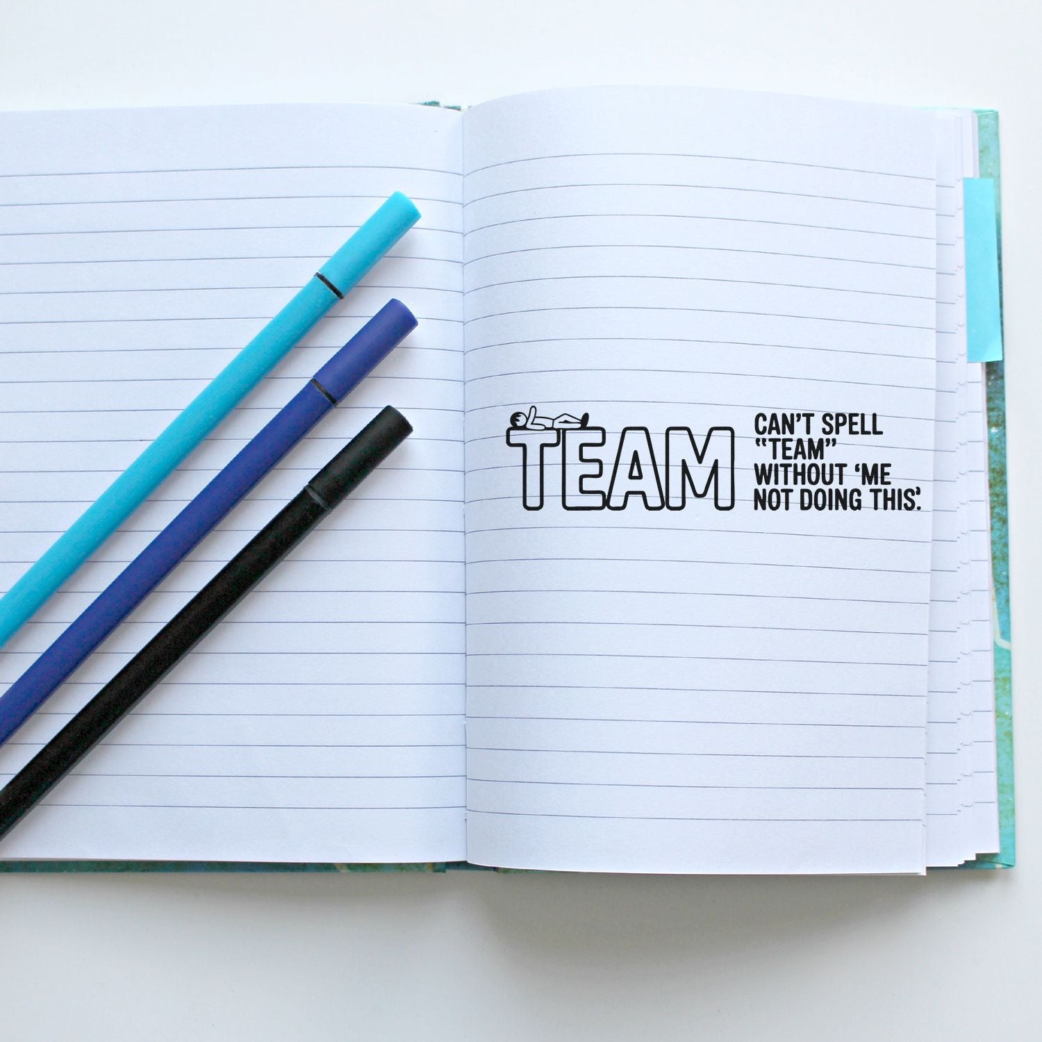 Open lined notebook with teal, purple, and black pens beside a black TEAM stamp reading Cant spell team without me.