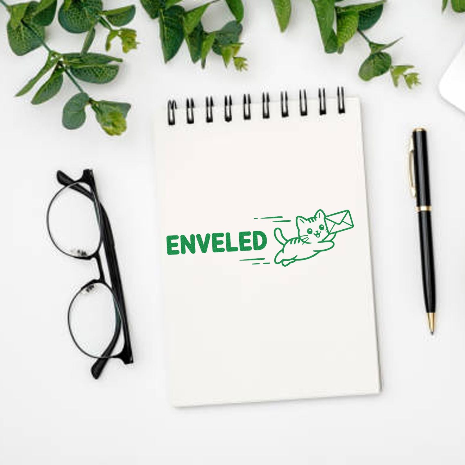 White notepad with green ENVELED cat-and-envelope stamp and seal design, beside black glasses and pen.