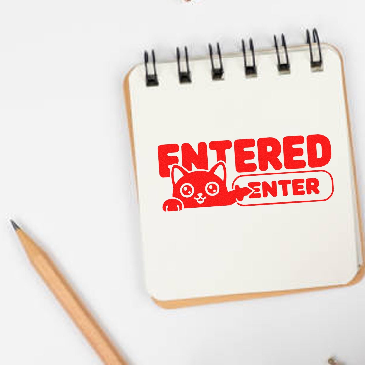 Red ENTERED stamp seal with cartoon cat on a white spiral notebook, beside a wooden pencil.