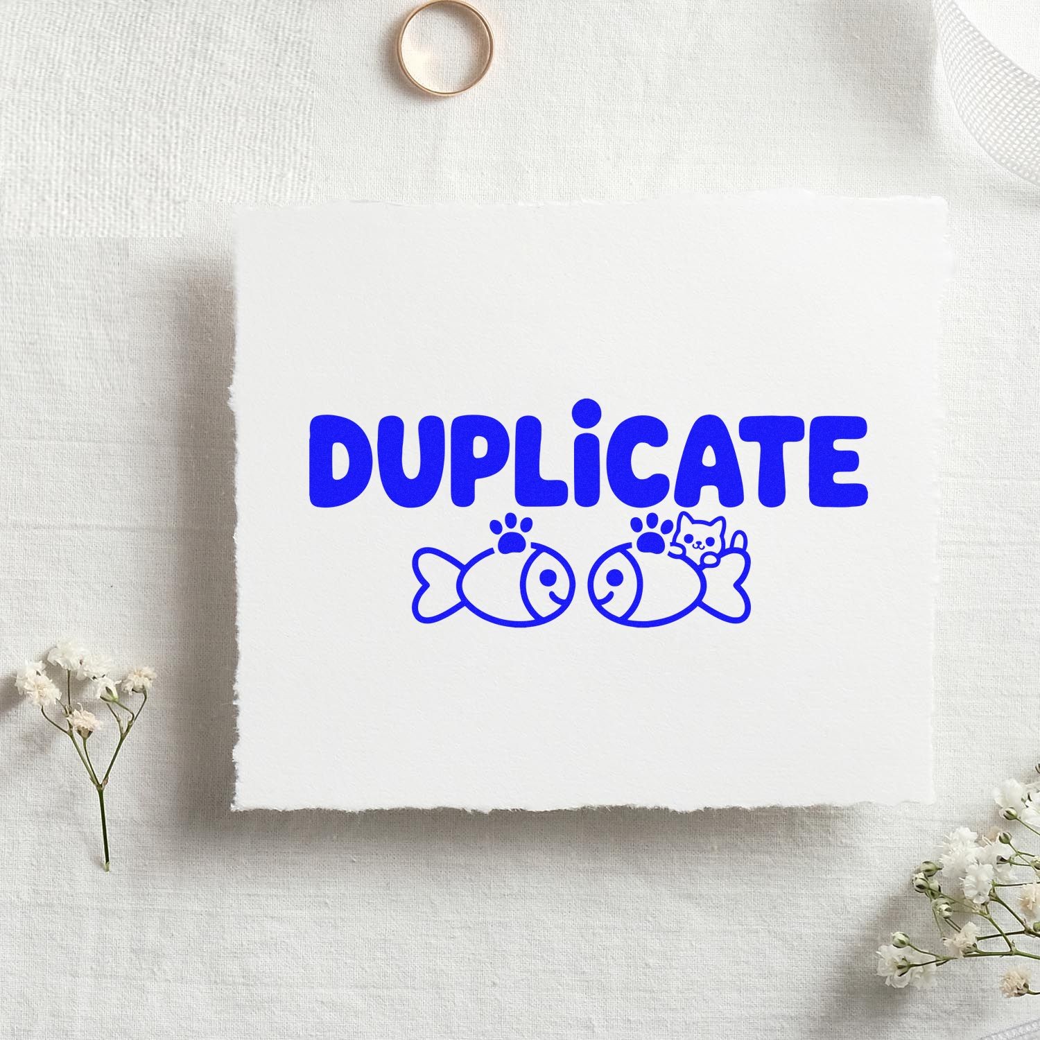 Blue DUPLICATE stamp and seal design with fish and cat motif on white handmade paper, on a soft white fabric background.