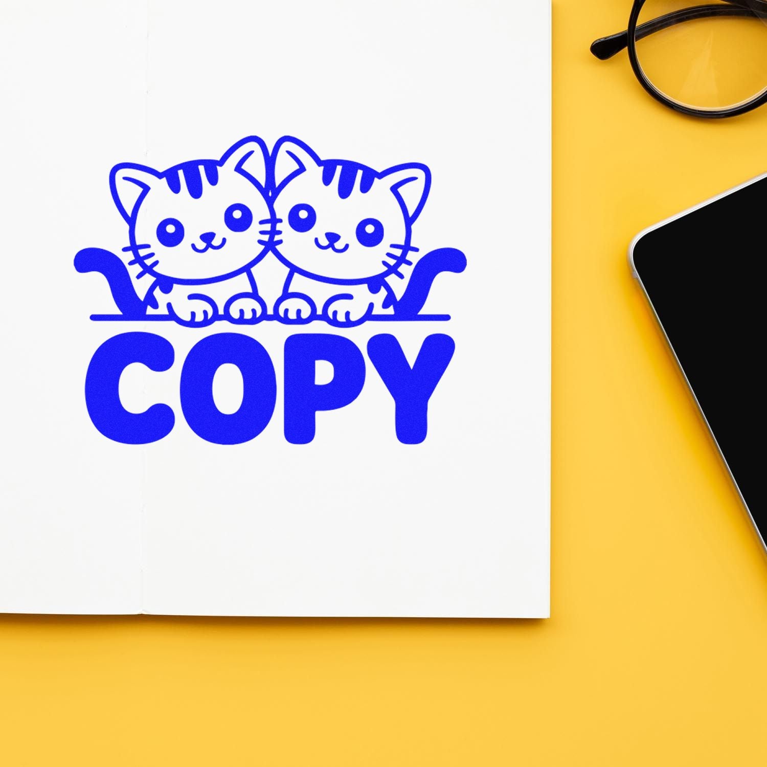 Blue cat COPY rubber stamp design on white page, yellow background, beside glasses and smartphone.