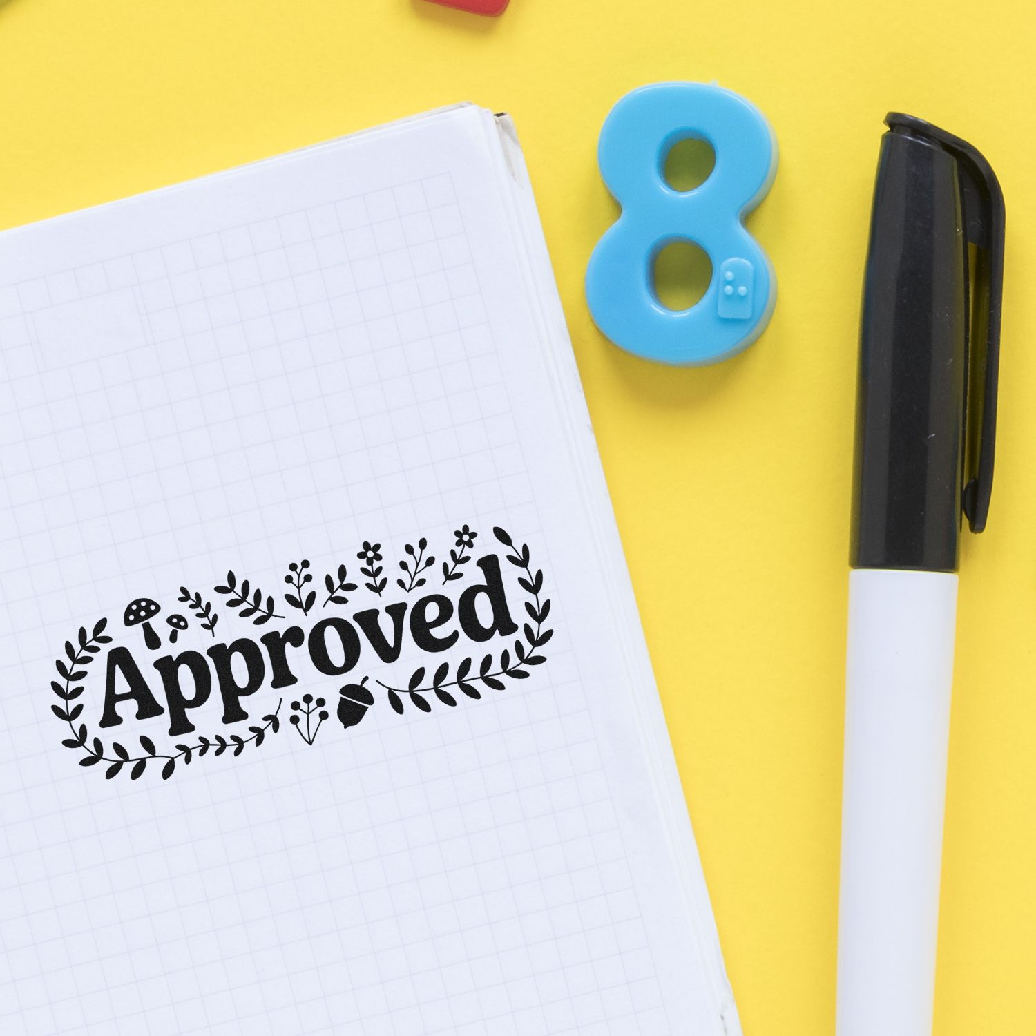 Black Approved stamp on grid notebook beside blue number 8 seal and black marker on yellow background.