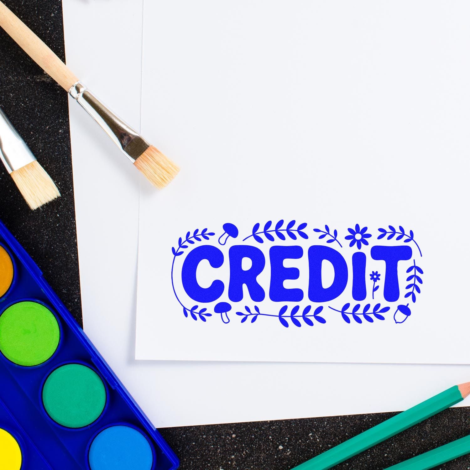 Blue decorative CREDIT stamp and seal on white paper with paint brushes, watercolor palette, and colored pencils