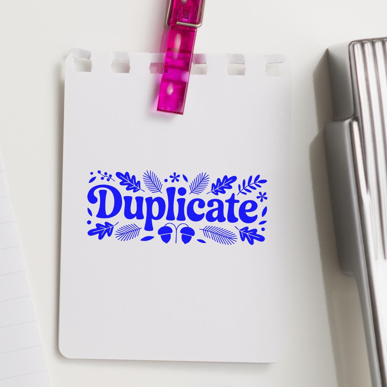 Blue decorative Duplicate stamp on white paper, clipped by pink translucent clip, with silver seal tool beside it.