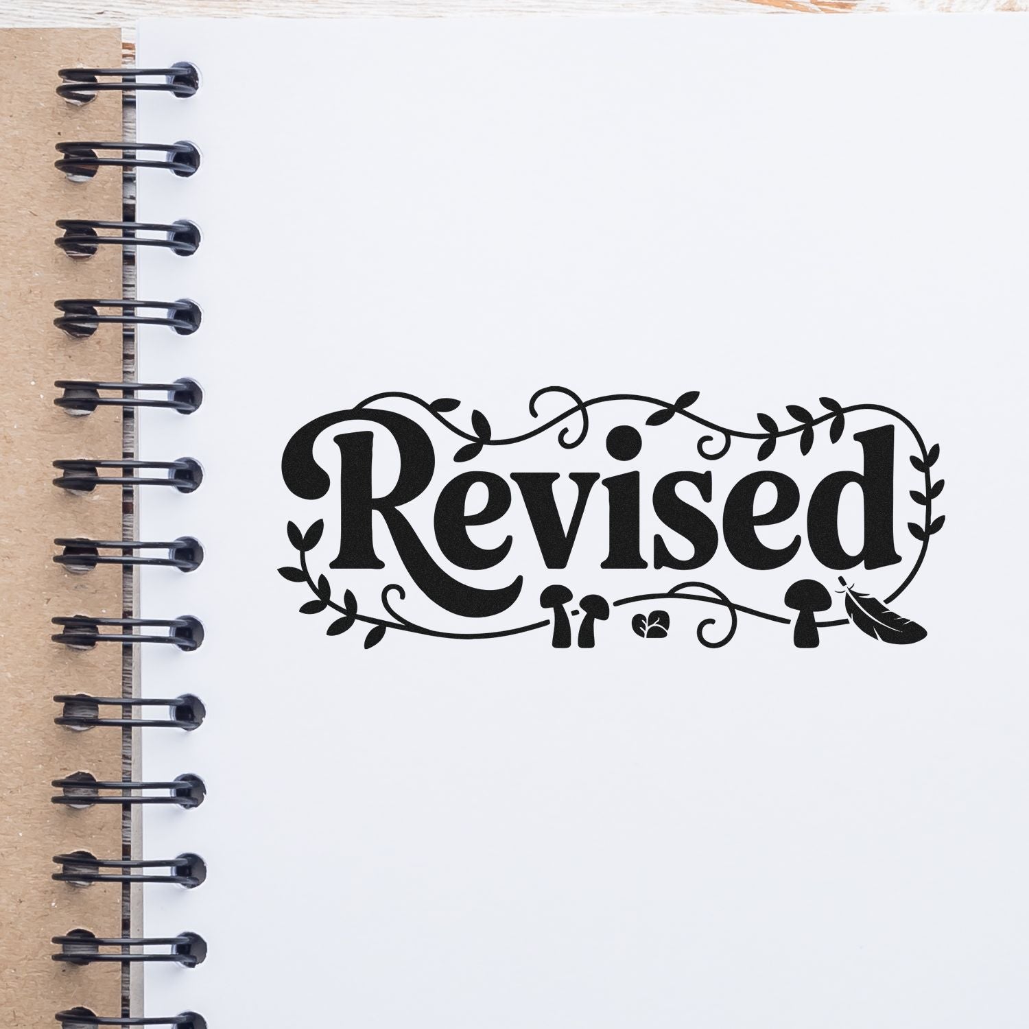 Black Revised stamp seal with vine flourishes on a white spiral notebook page and tan cover.