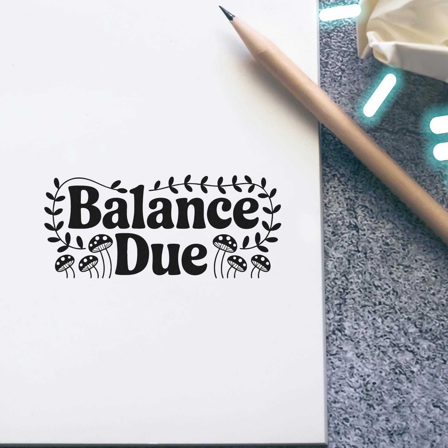 Black Balance Due stamp with leafy and mushroom seal design on white paper beside a pencil, on a gray textured surface.