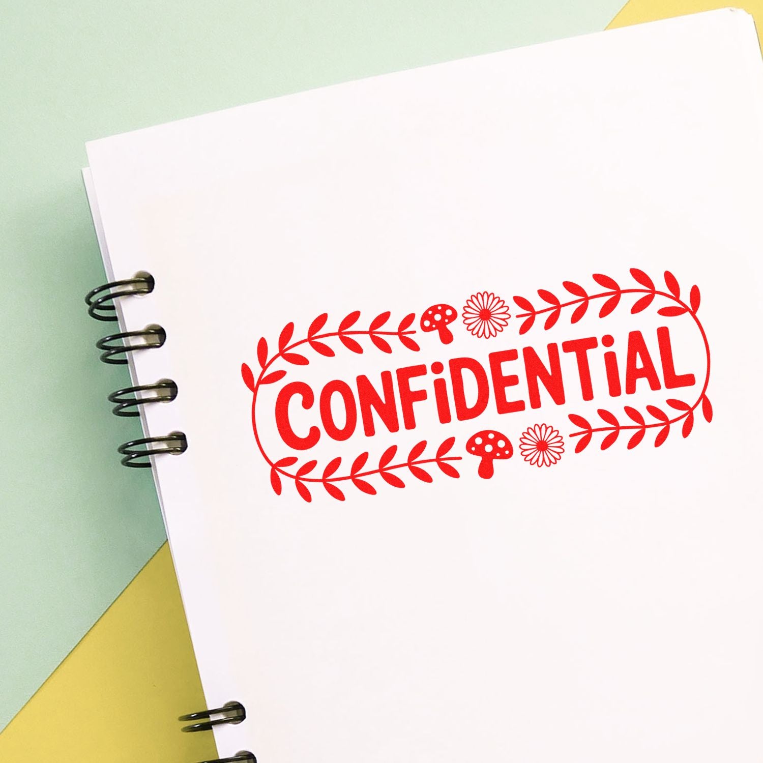 Red CONFIDENTIAL stamp seal with leaf border and mushroom motifs on a white spiral notebook, over soft green and yellow paper.