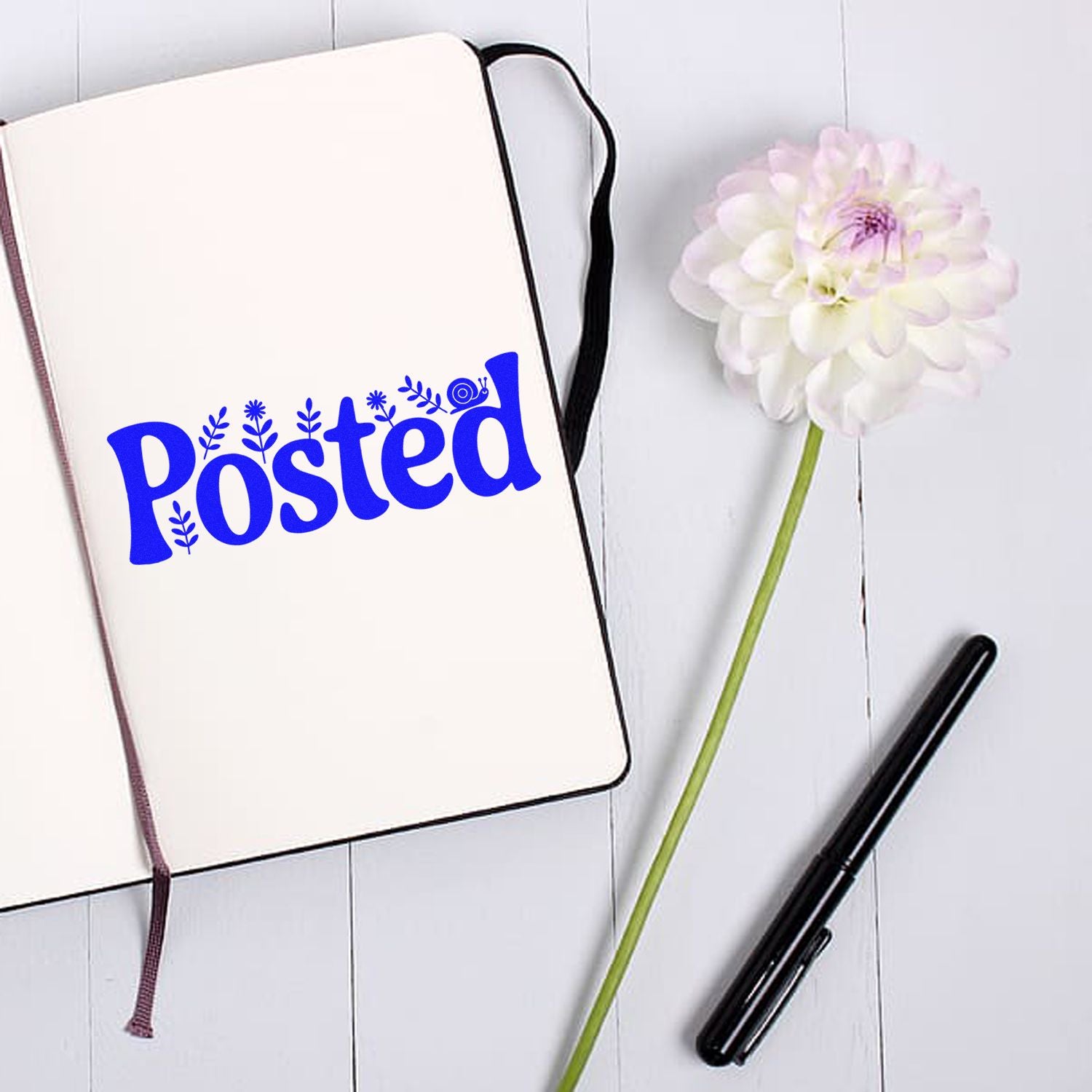Blue Posted stamp and seal on a cream notebook beside a white flower and black pen.