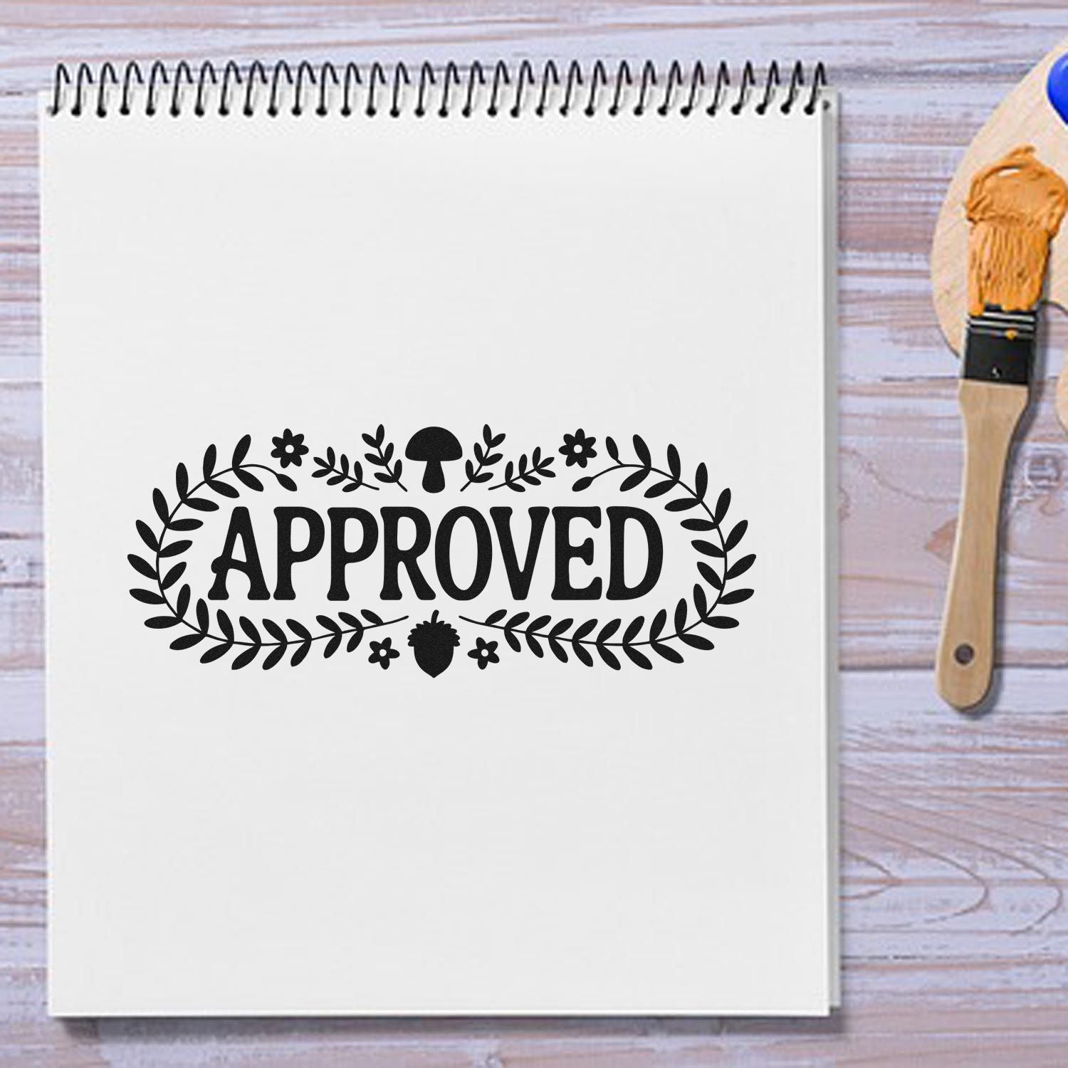 Black APPROVED rubber stamp seal with leaf wreath on a white sketchbook, beside a paintbrush on a wood table.