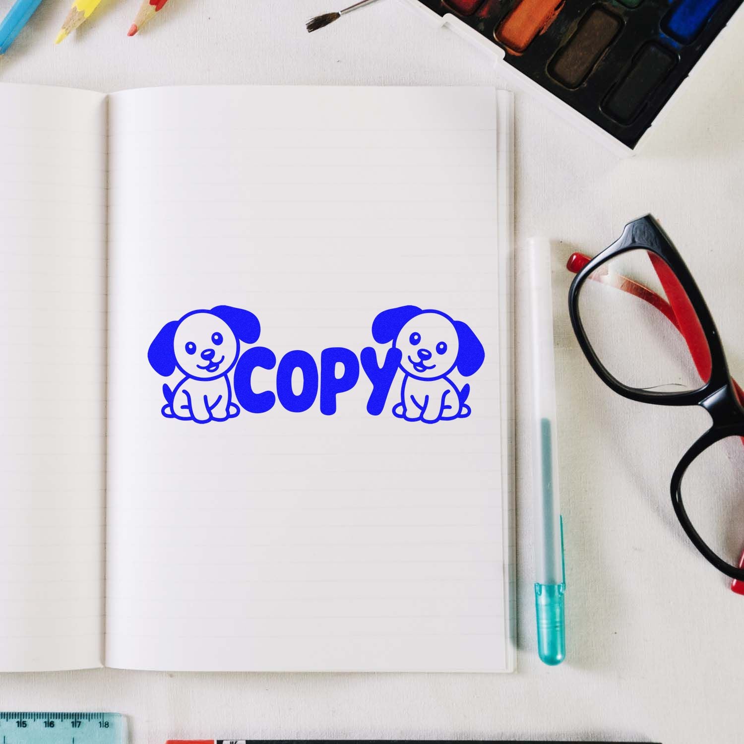 Blue COPY stamp with two cartoon dog seal designs on a lined notebook, beside glasses, pen, and watercolor tools.