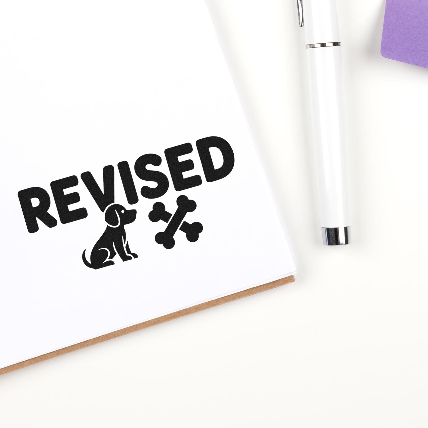 Black REVISED stamp and dog-bone seal beside a white pen on a white desk