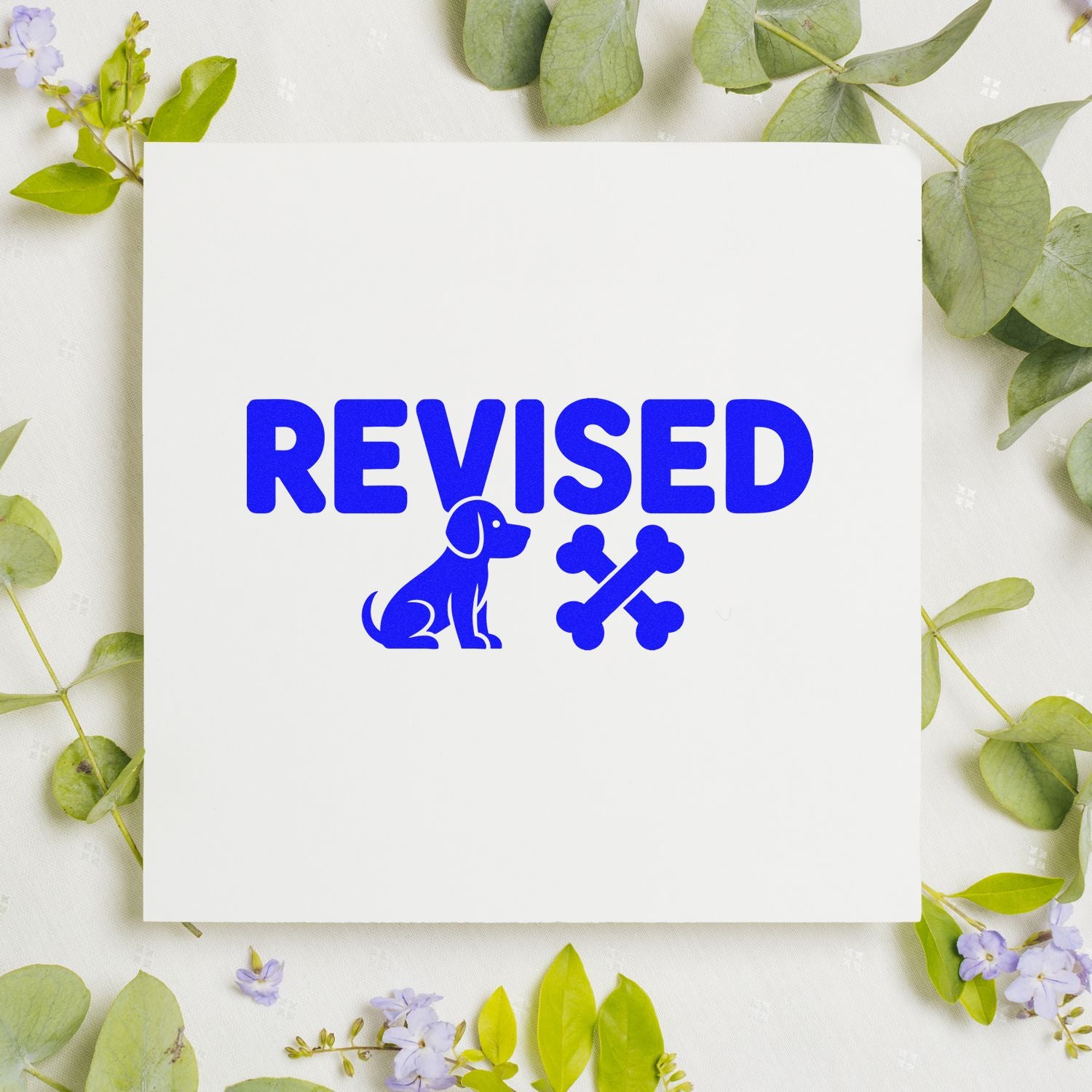 Blue REVISED dog and bone stamp/seal on white card, framed by green leaves and lavender flowers.