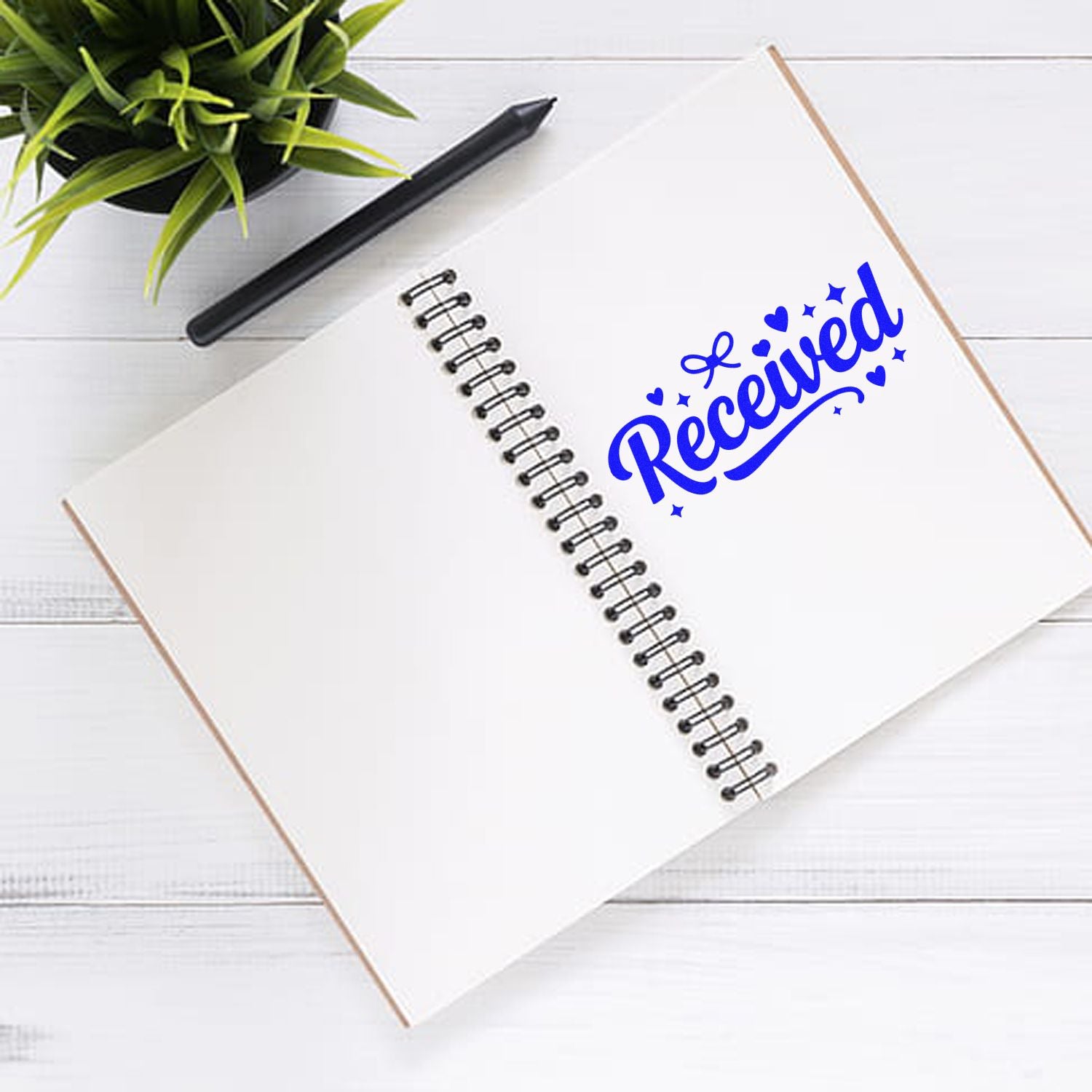 Blue Received stamp seal on an open spiral notebook, with a black pen and green plant on a white desk.