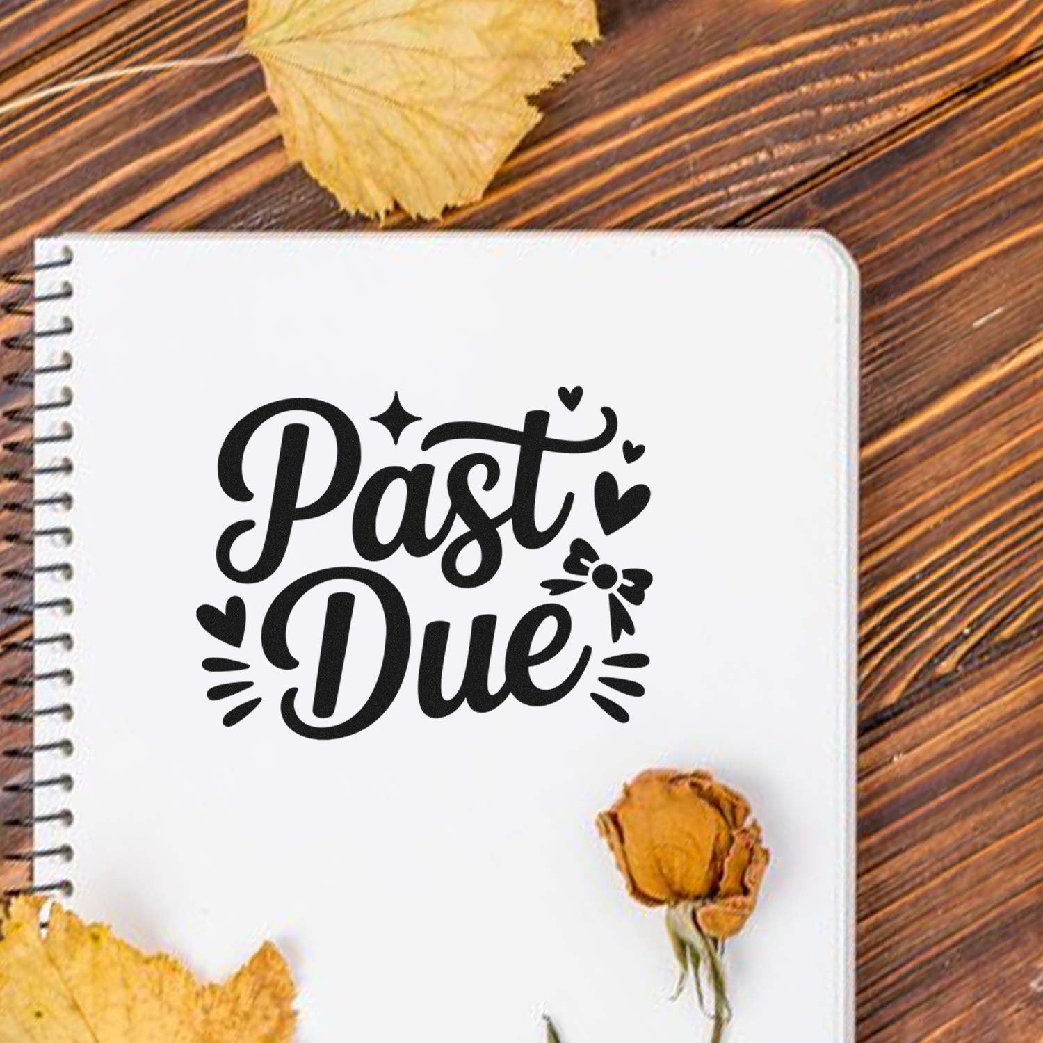 Black Past Due stamp/seal design on a white spiral notebook, with heart accents on rustic wood and yellow leaves.